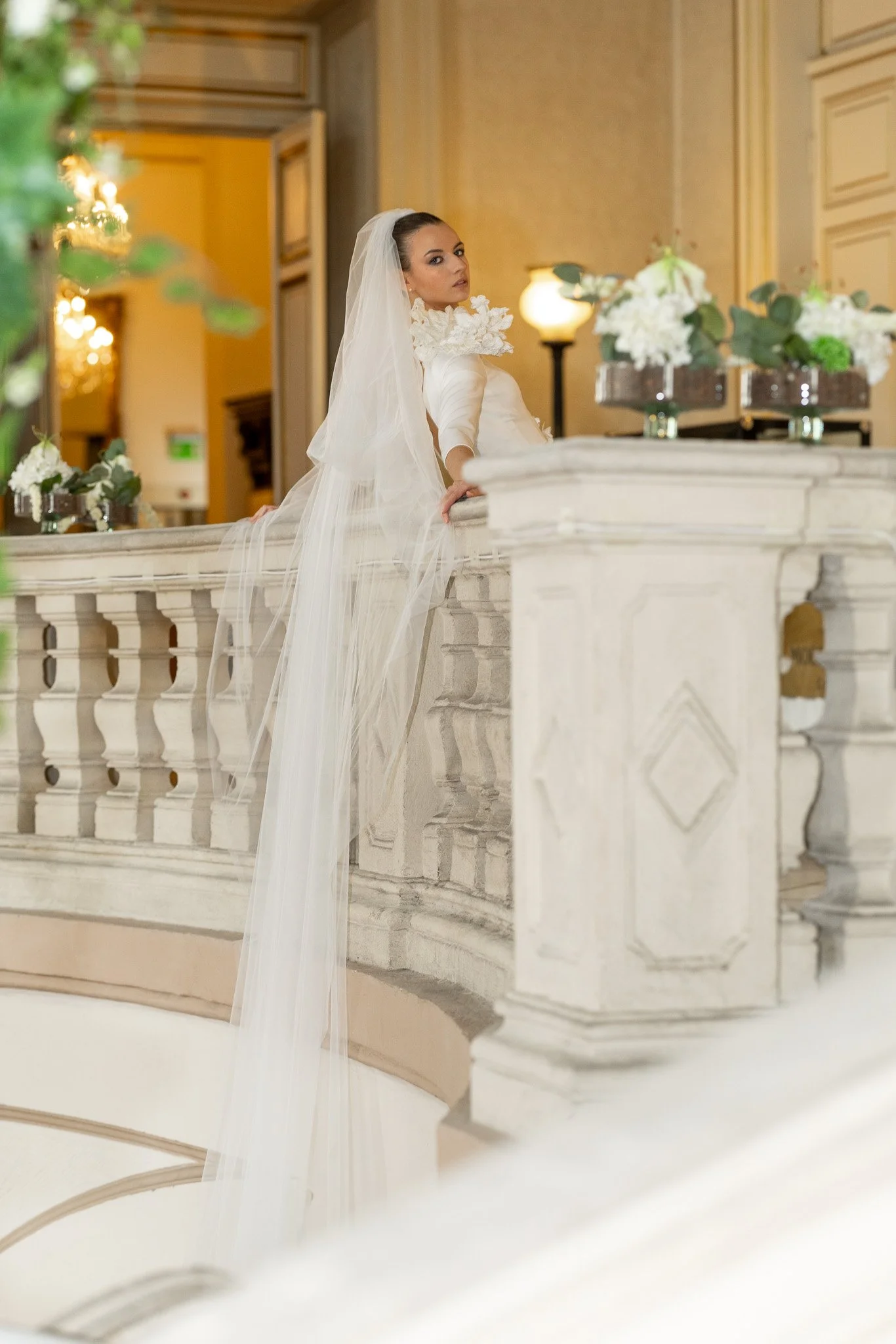 A woman dressed in a white wedding gown with a long veil, leaning on a marble railing in an elegant, historic building with floral arrangements and warm lighting.