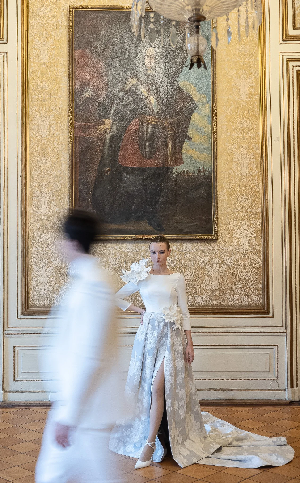 A woman in a white floral gown standing in front of a large framed portrait in an ornate room.