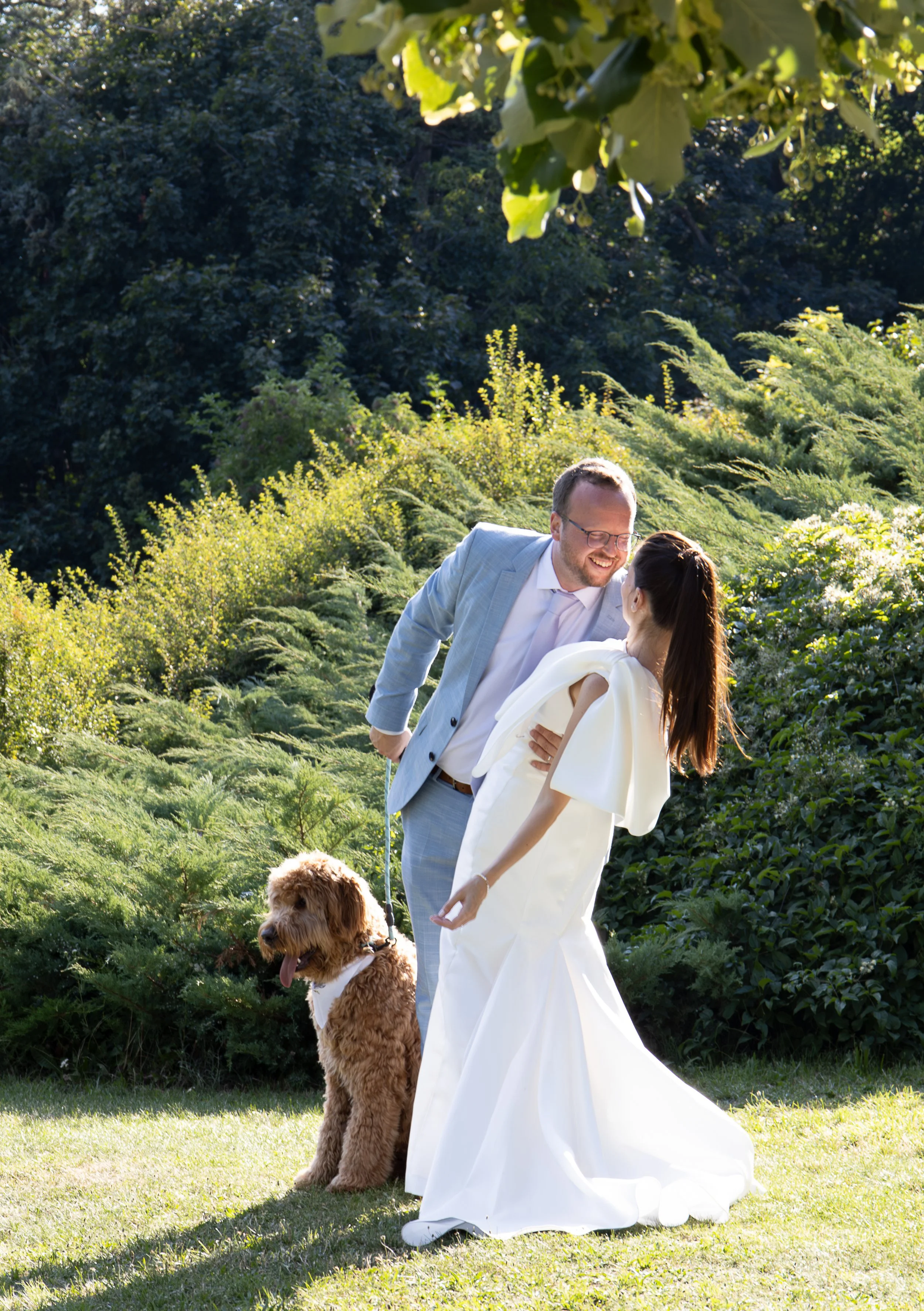 A couple in wedding attire with a dog outdoors on a sunny day, surrounded by greenery.