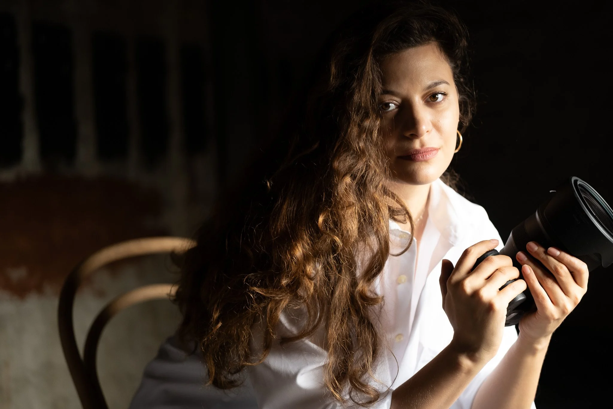 A woman with long, wavy brown hair, wearing a white shirt, holding a camera and looking at the viewer, in a dimly lit environment.