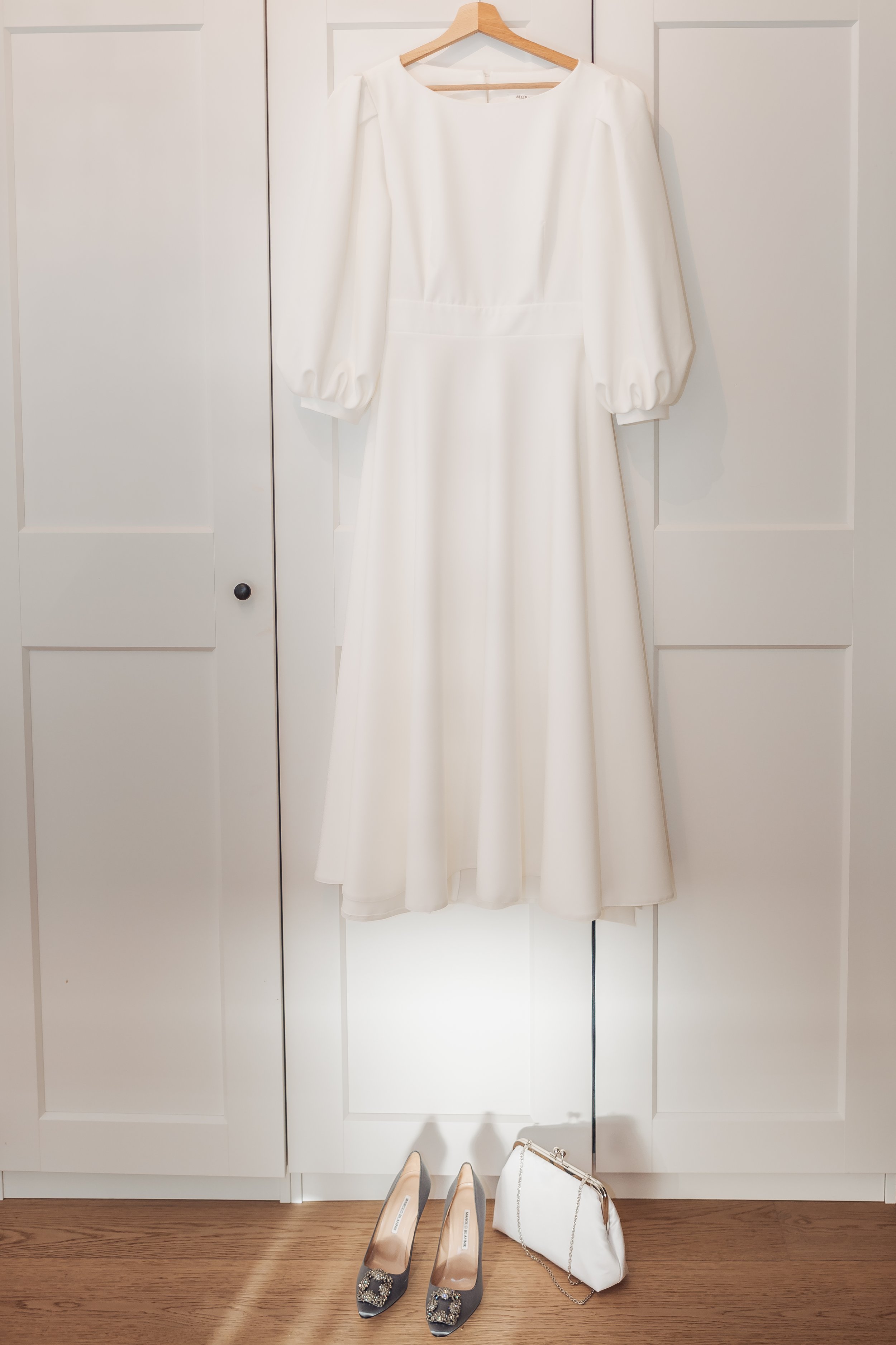 A white dress hanging on a hanger on a door, with a pair of gray high heels with embellishments and a white purse with a chain strap placed on the floor.