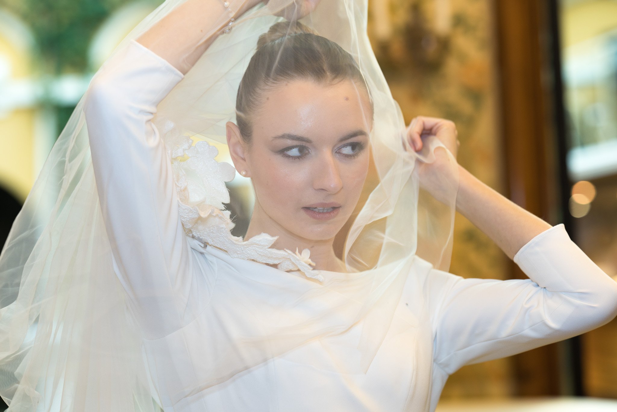 A Wedding Photographer in Palais Auersperg Vienna.  bride wearing a white dress adjusting a sheer veil on her head, with a blurred background of wooden and green elements.