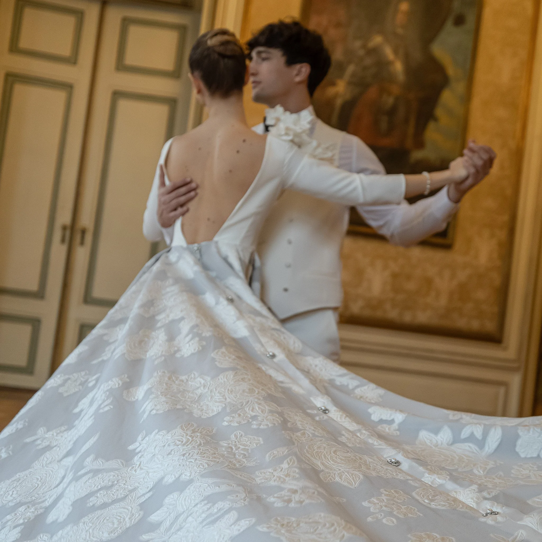 Two individuals dancing closely, with one wearing a white dress with lace details and the other in a white shirt with a large floral accessory. They are indoors, in a room with a wooden floor and a wall with painted wooden panels and a large framed p