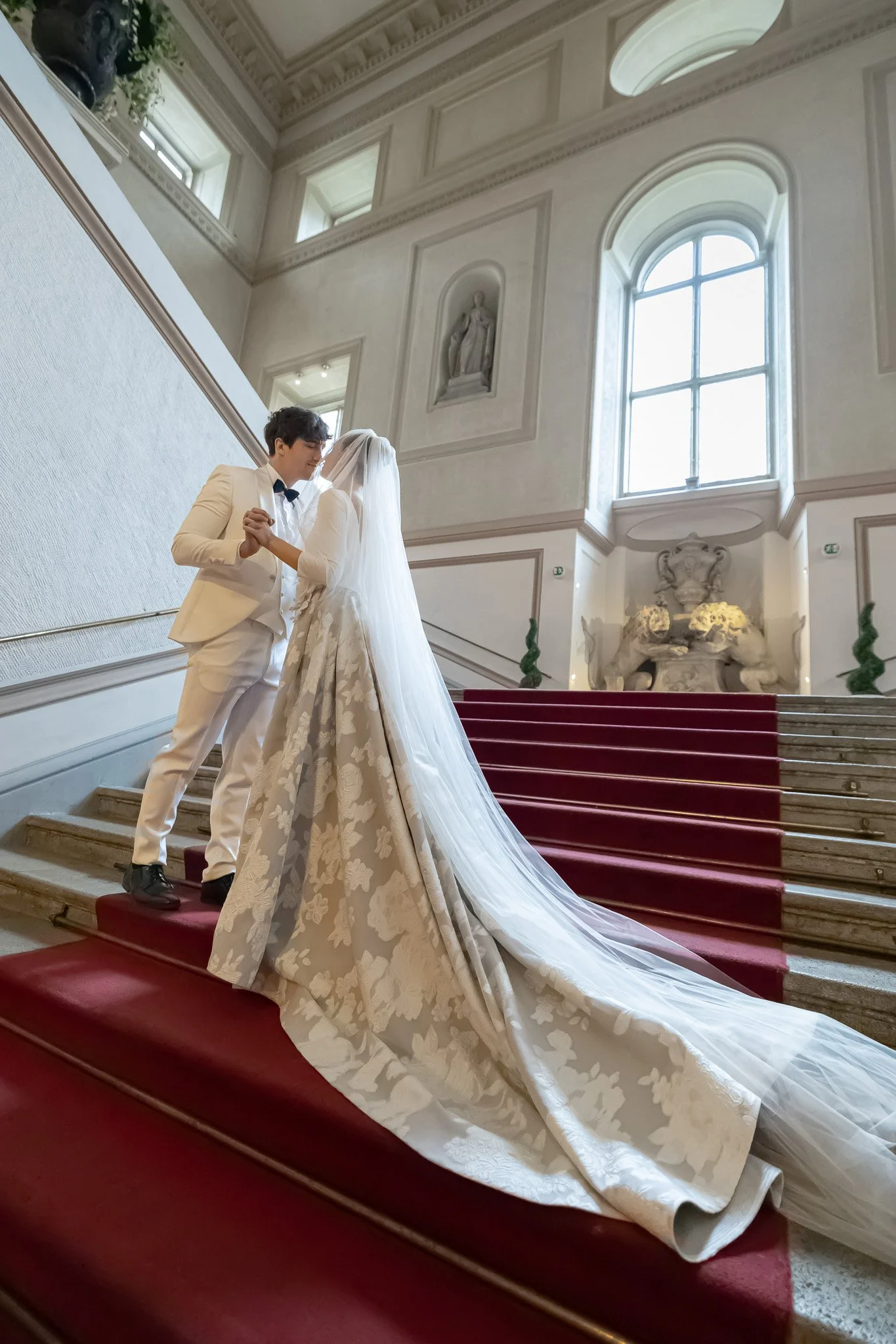 Wedding Photography in Palais Auersperg Vienna. A bride and groom dance on a red-carpeted staircase inside a grand, ornate building, with large windows and decorative statues and sculptures in the background.