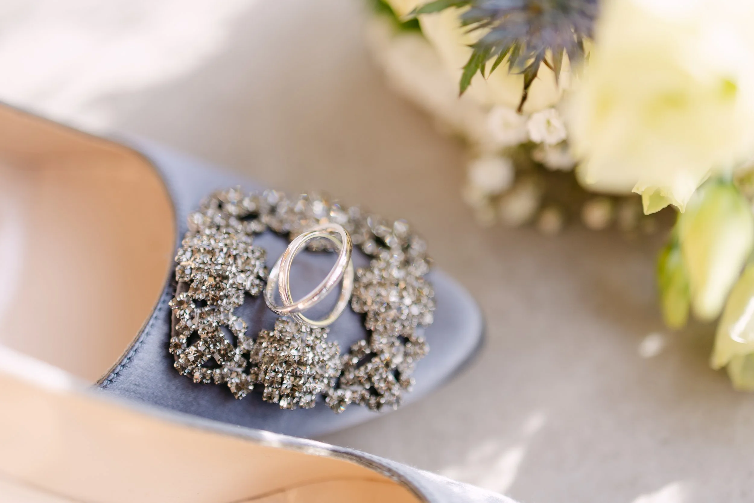 Two wedding rings resting on a decorative, glittering silver brooch inside a light-colored jewelry box, with blurred flowers in the background.