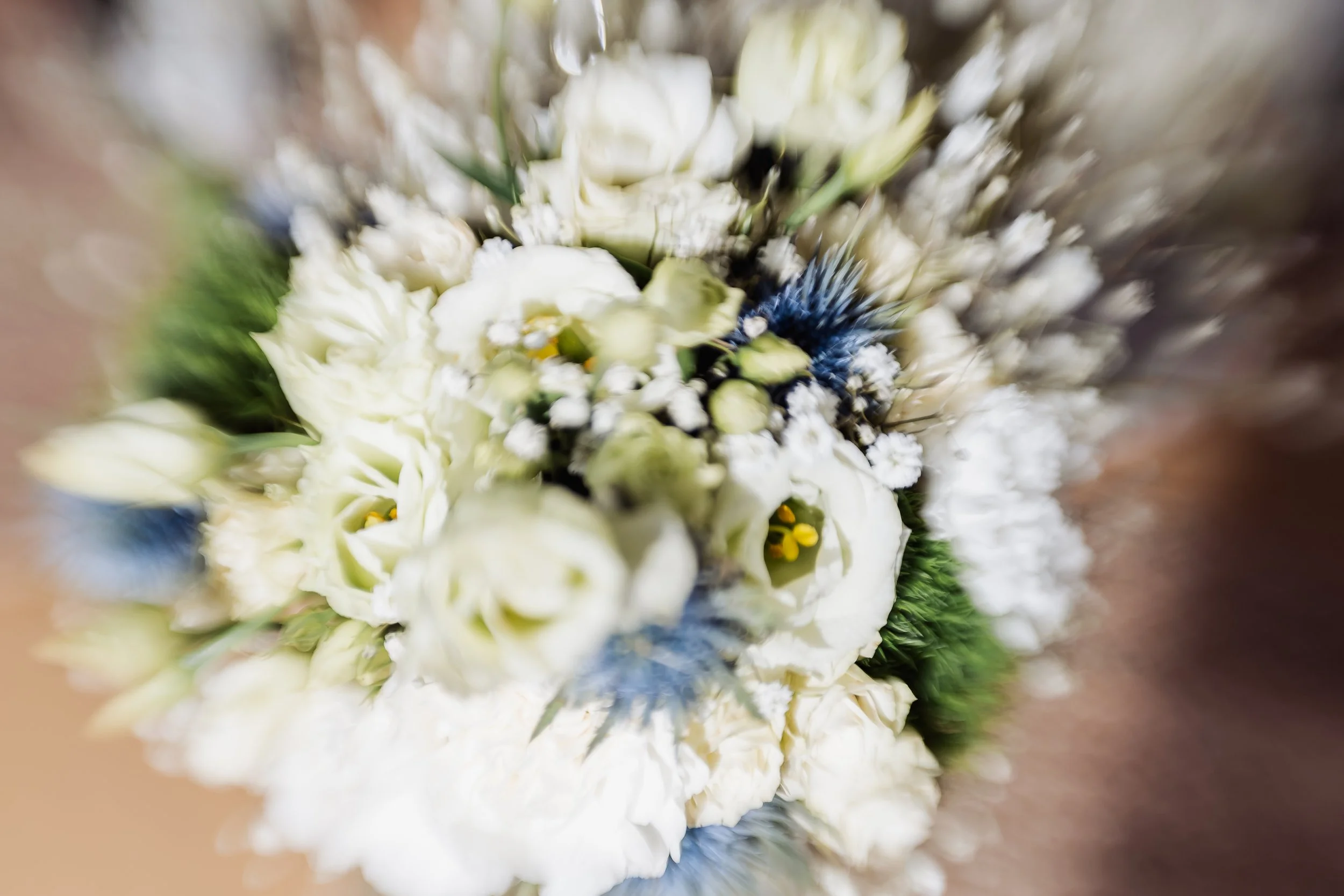 A blurred close-up of a bouquet of white flowers, including ranunculus, baby's breath, and blue thistle.