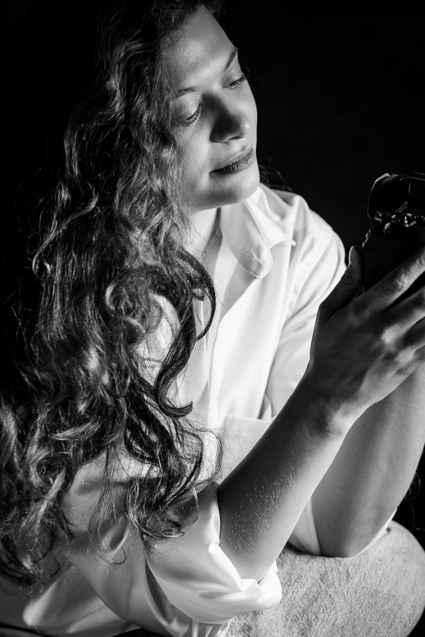 A woman with long, curly hair wearing a white shirt, looking at a camera in her hand in a black and white photo.