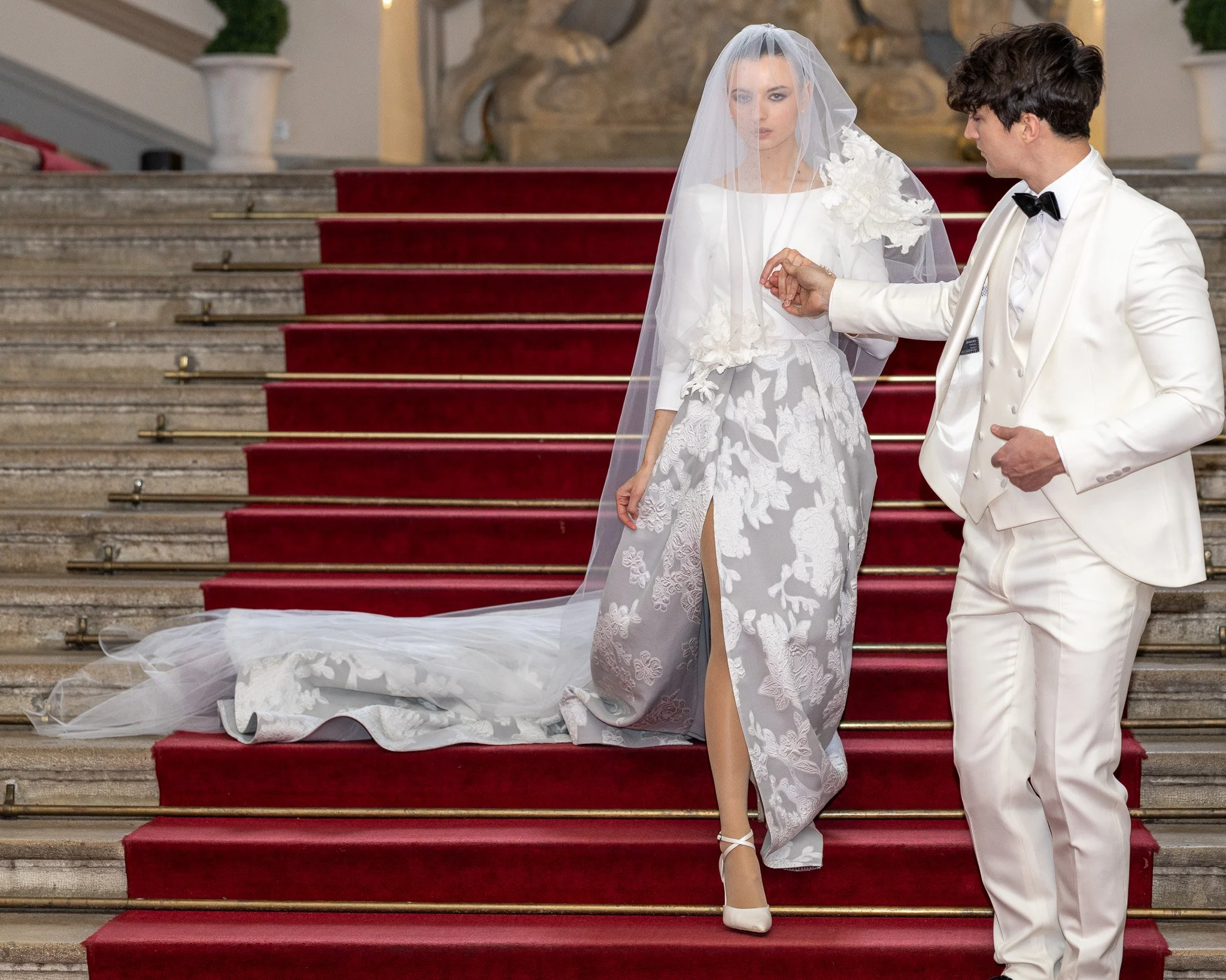 A bride and groom stand on red-carpeted stairs during their wedding ceremony inside a building with stone walls and ornate decorations. The bride is wearing a white lace wedding dress with floral details, a veil, and high heels. The groom is in a whi