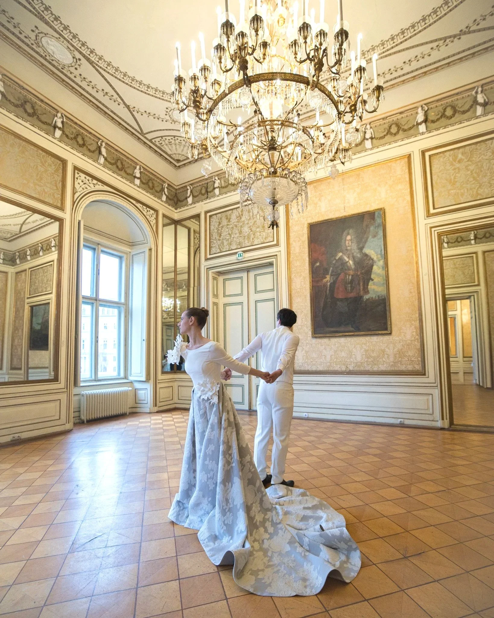 A bride and groom holding hands in a grand, elegant hall with ornate gold and cream decor, high windows, and a large chandelier hanging from the ceiling.