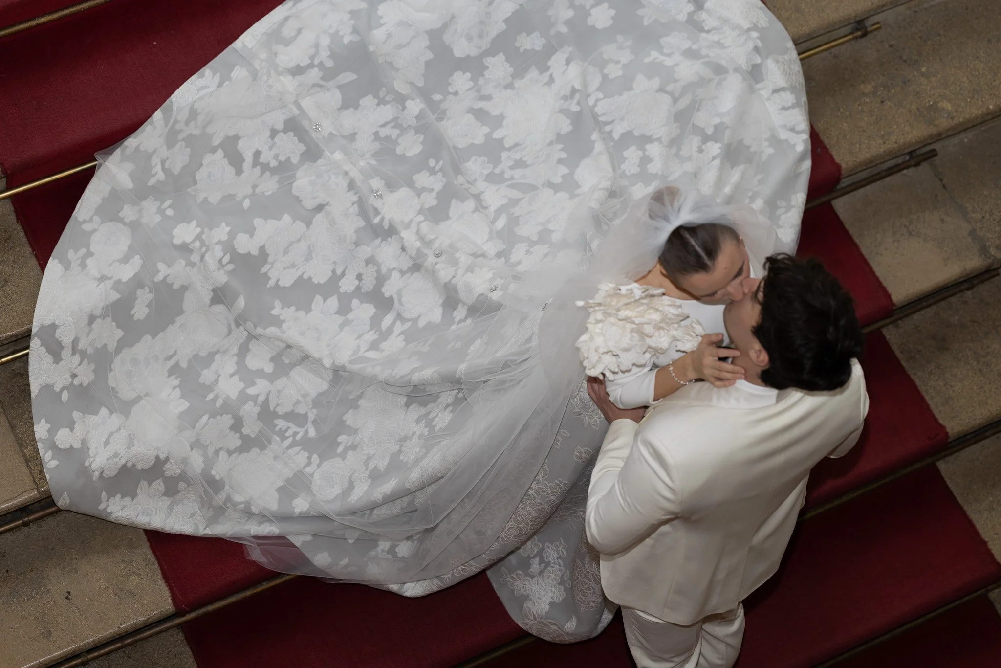 A bride and groom kiss during their wedding ceremony in a church, seen from above. The bride wears a white gown with floral embroidery and a veil; the groom is in a white suit.