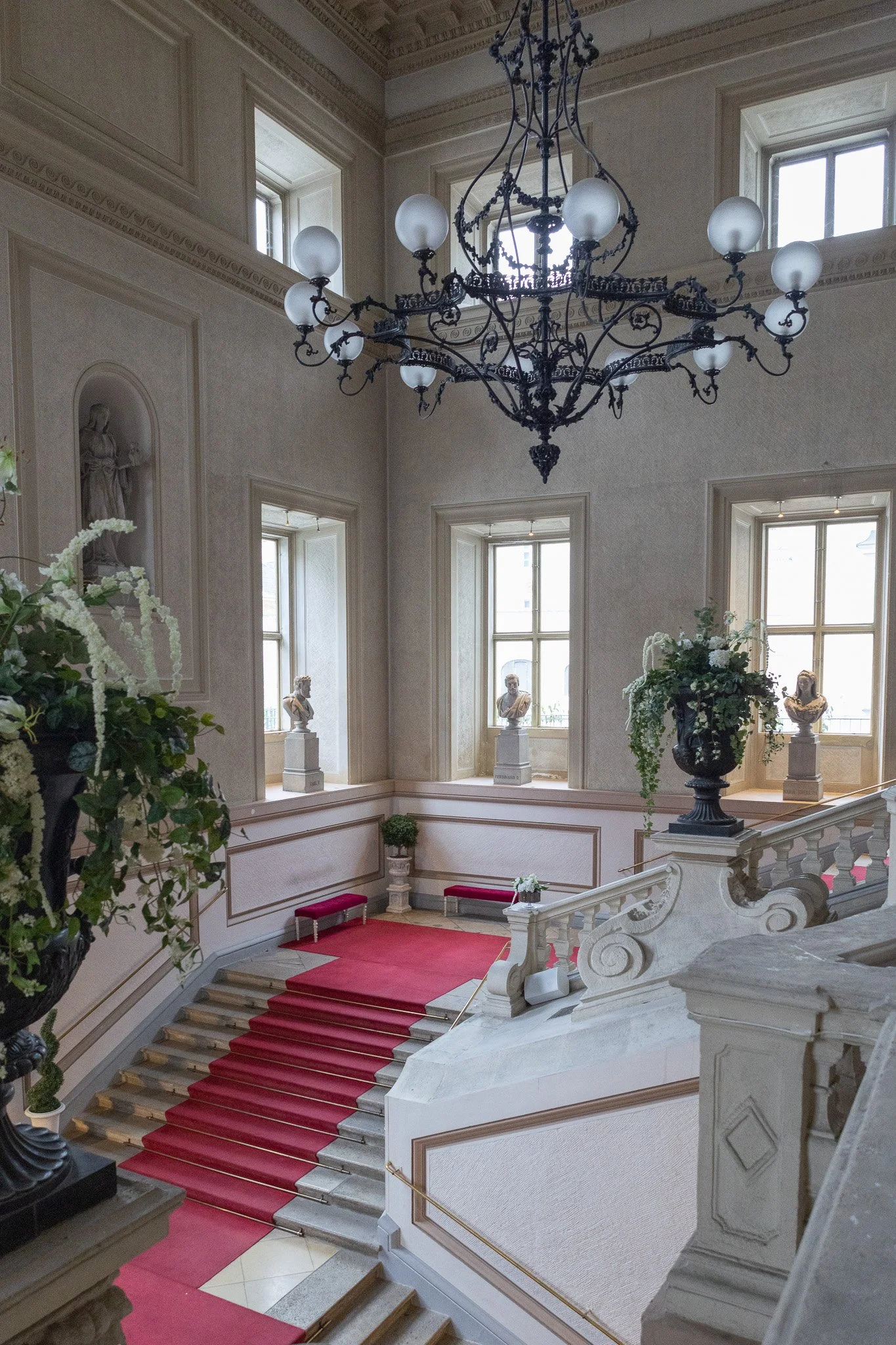 Elegant staircase with a red carpet runner, ornate white balustrade, and large black chandelier in a luxurious interior, adorned with bust sculptures and floral arrangements.
