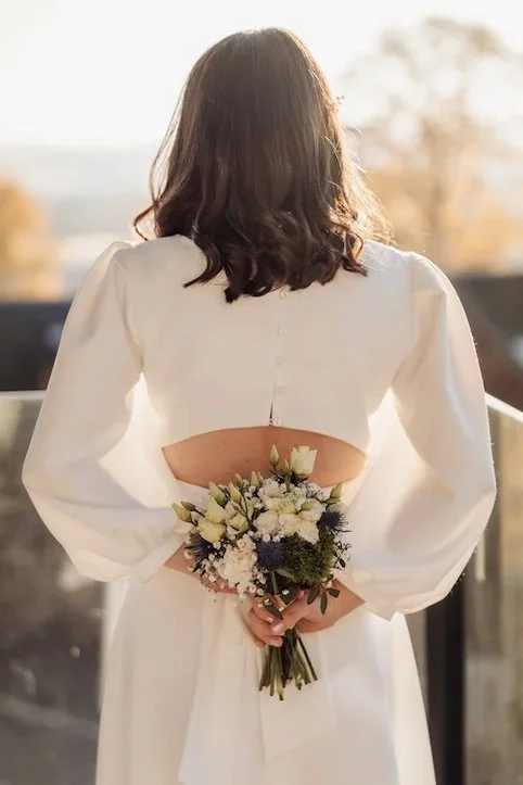 Woman in white dress holding a bouquet of white and purple flowers behind her back, facing away outside during sunset.