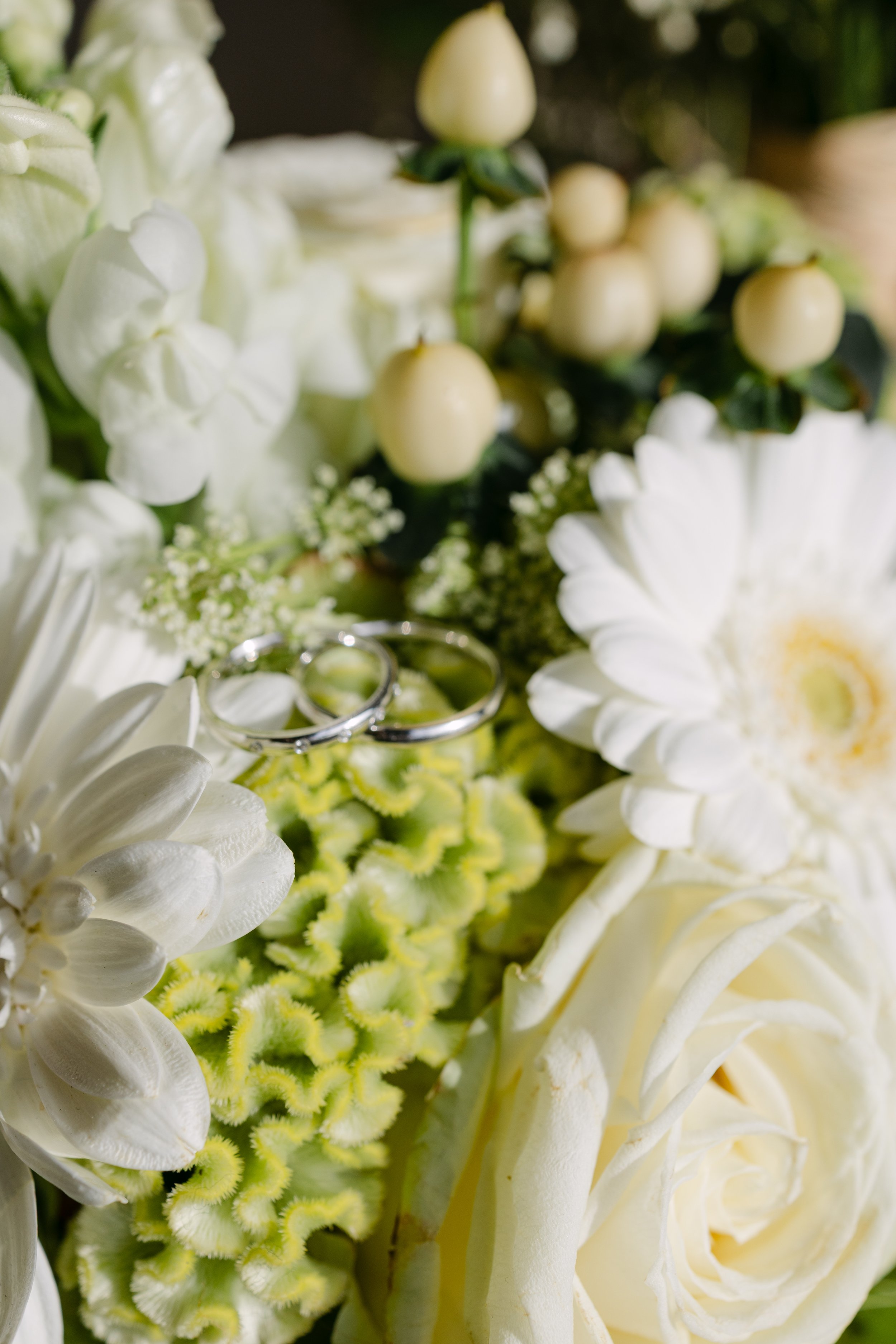 Close-up of a wedding bouquet with white roses, green flowers, and white berries, with two silver rings resting on the flowers.
