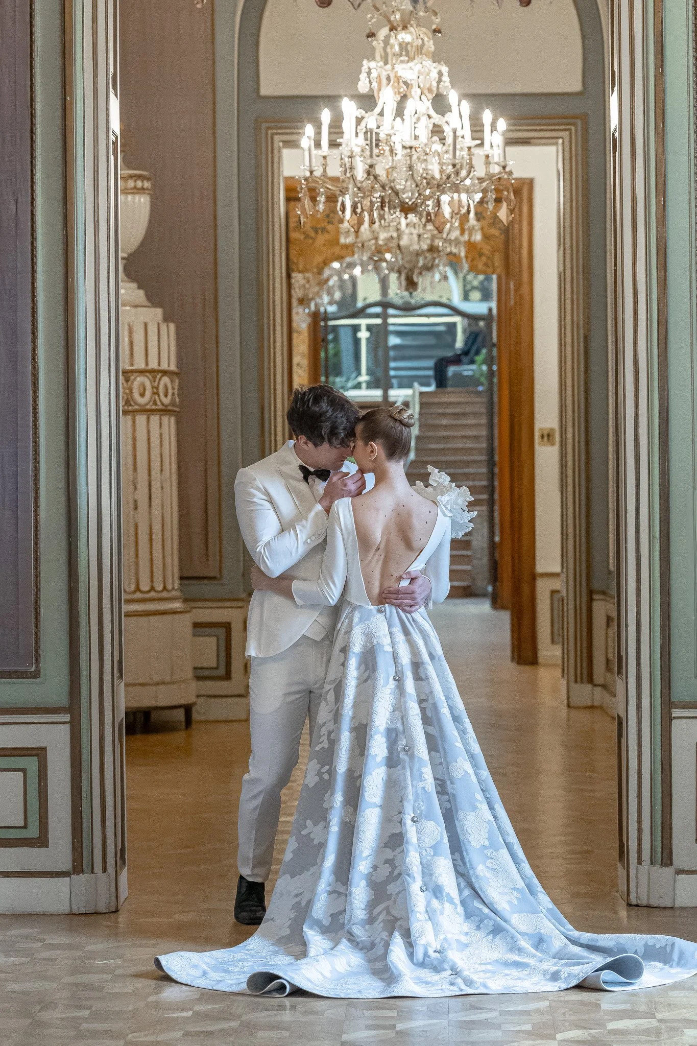 A couple in wedding attire sharing a tender moment in an elegant, ornate hallway with a large chandelier hanging from the ceiling.