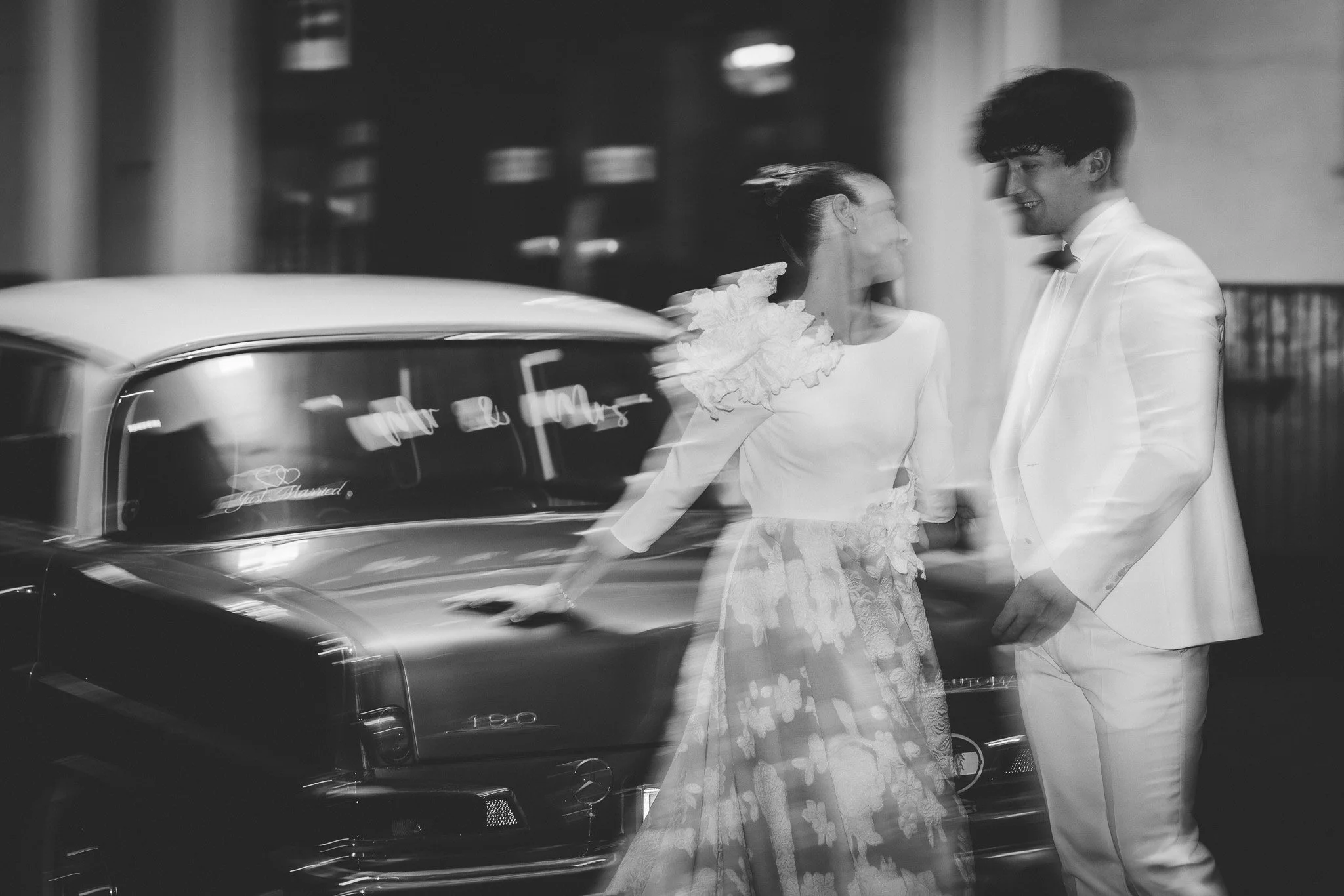 A black and white photo of a bride and groom standing beside a classic car, looking at each other and smiling. The bride is wearing a dress with ruffled sleeves and a floral pattern, and the groom is dressed in a suit with a bow tie. The photo captur