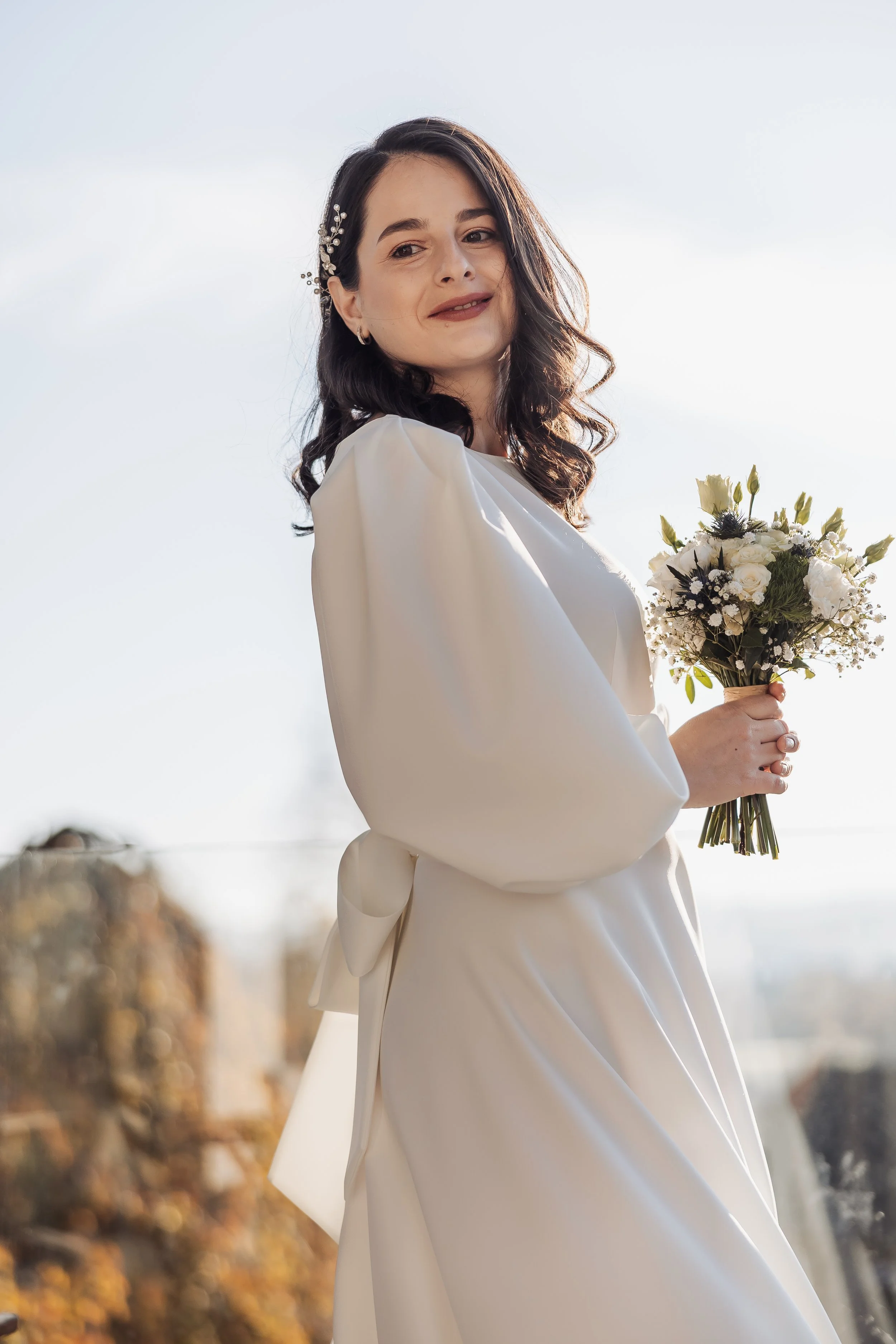 A woman in a white dress holding a bouquet of white and green flowers, standing outdoors with a bright sky and blurred autumn trees in the background.