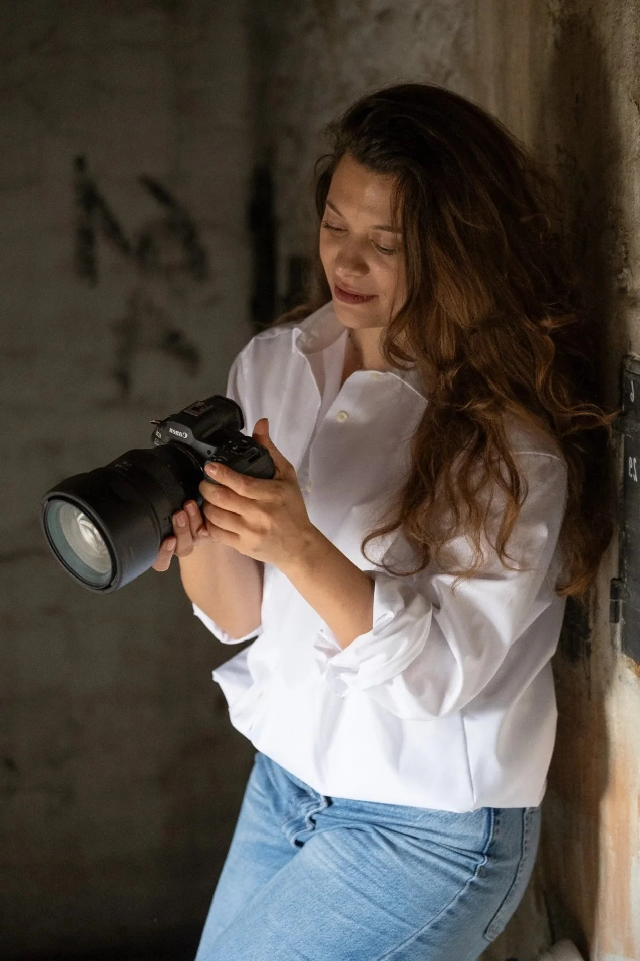A young woman with long wavy brown hair wearing a white shirt and blue jeans, leaning against a brick wall, while holding and looking at a professional camera.