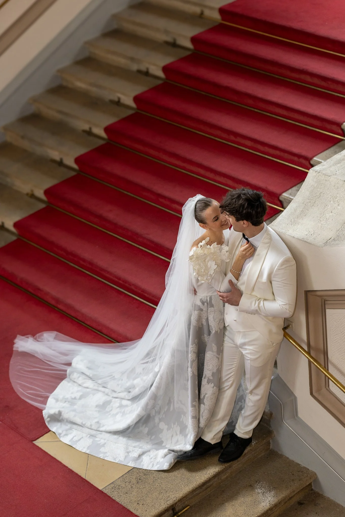 A bride and groom are standing on a staircase with red carpet, smiling and looking at each other, with the bride wearing a white wedding gown with a long train and veil, and the groom in a white tuxedo with black shoes.