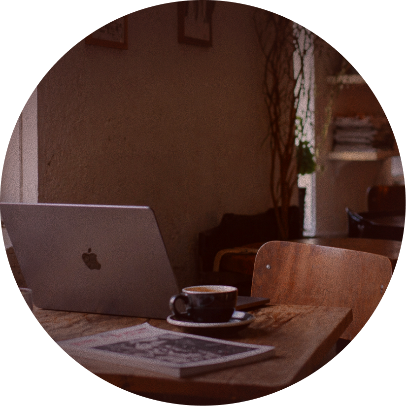 A cozy cafe setting with a wooden table holding a silver MacBook, a black coffee cup with a saucer, and a magazine. In the background, there are shelves with books and decorative plants, and a window letting in natural light.