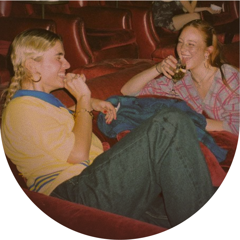Two women sitting on theater seats, smiling and laughing, one drinking a glass of wine.