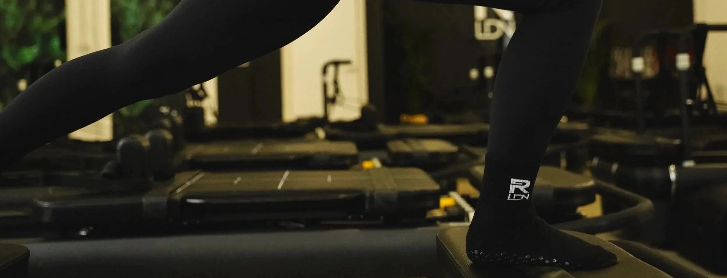 Close-up of a person in black leggings and socks with white text, standing on a step in a gym with workout equipment in the background.