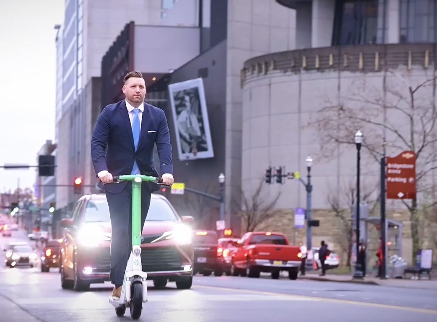A man in a business suit riding a green electric scooter across a city street with cars and city buildings in the background.