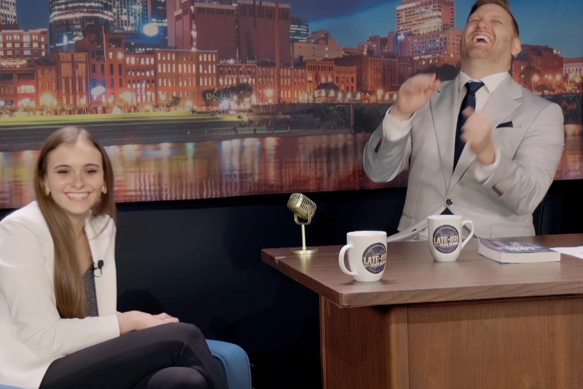 A late night host and his guest are laughing during a talk show, with a cityscape backdrop, a microphone, and two coffee mugs on the desk.