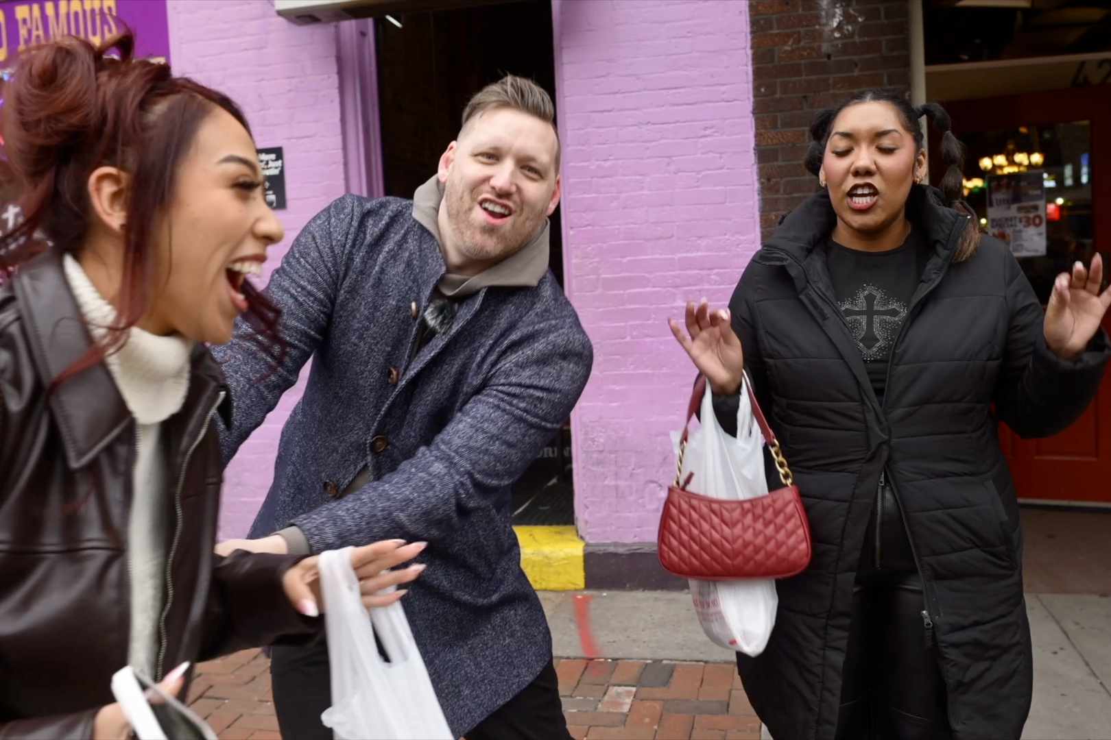 Three people outside, two women and one man, appear to be in a lively conversation or argument, with animated facial expressions. The women are holding shopping bags and wearing black jackets, while the man is wearing a dark coat over a hoodie. They are standing in front of a pink and brick building.