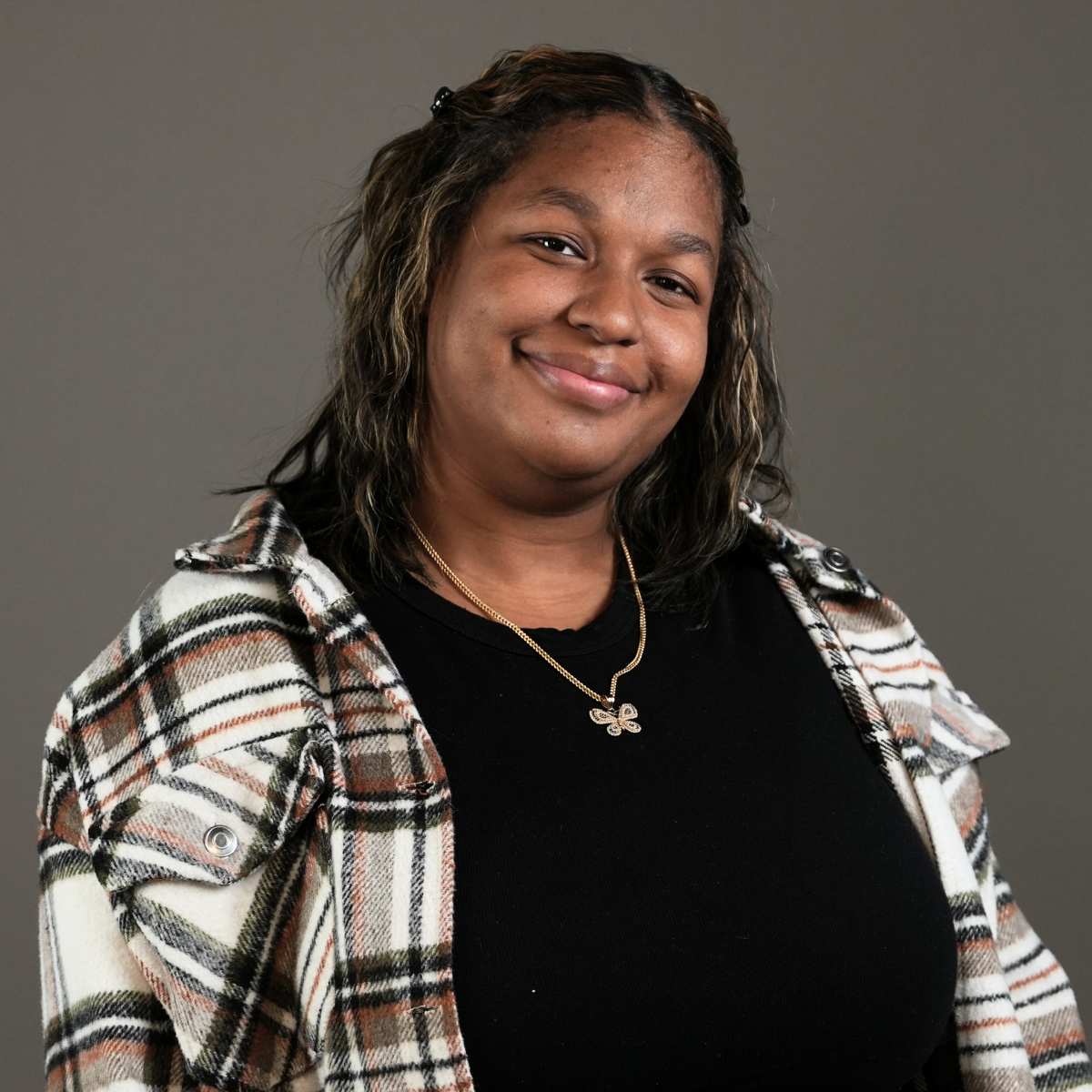 A young woman with curly black hair, glasses, earrings, and necklaces, smiling in a room with blurred background.