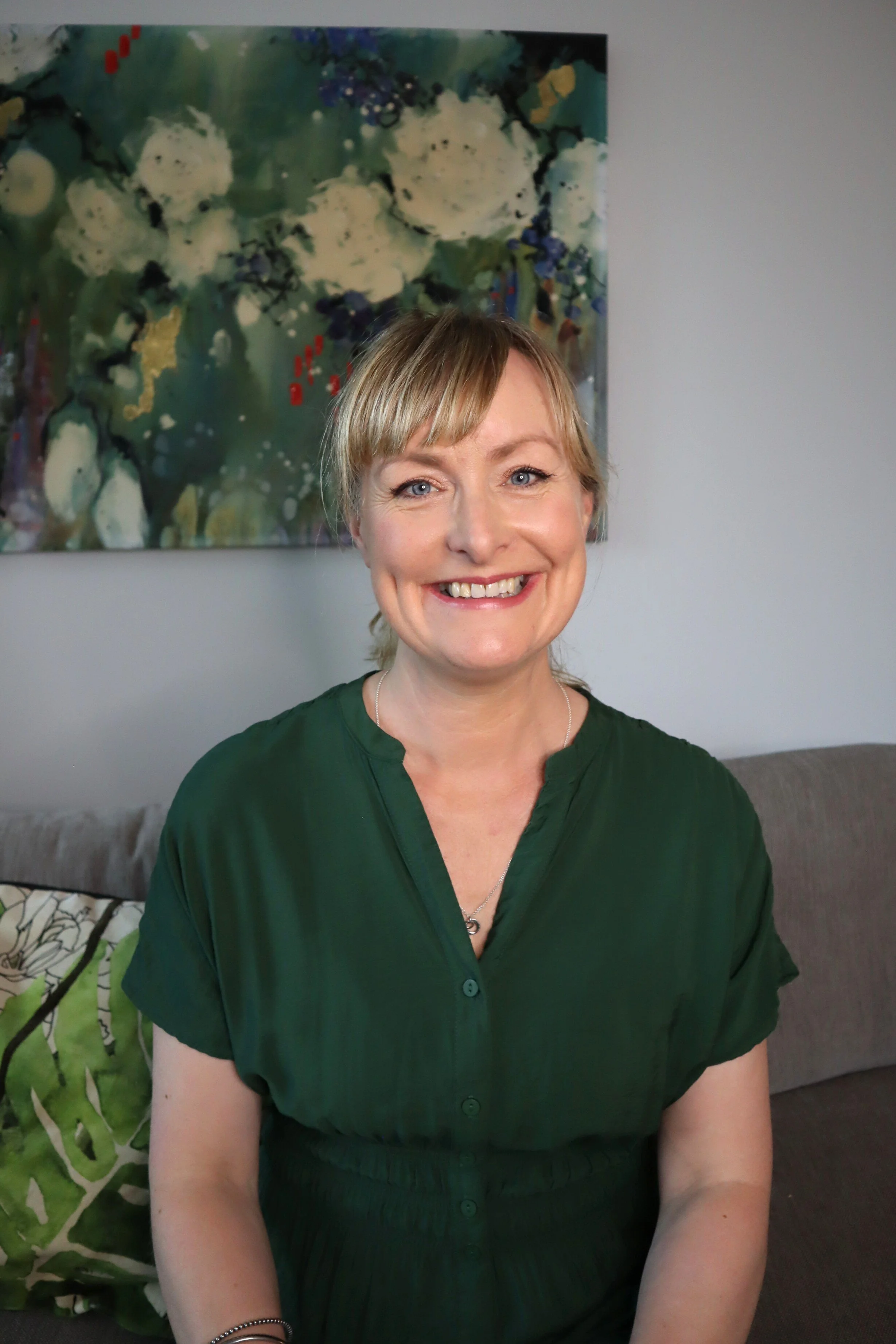 A woman sitting on a grey sofa in a living room, smiling, with a colourful abstract painting on the wall behind her.