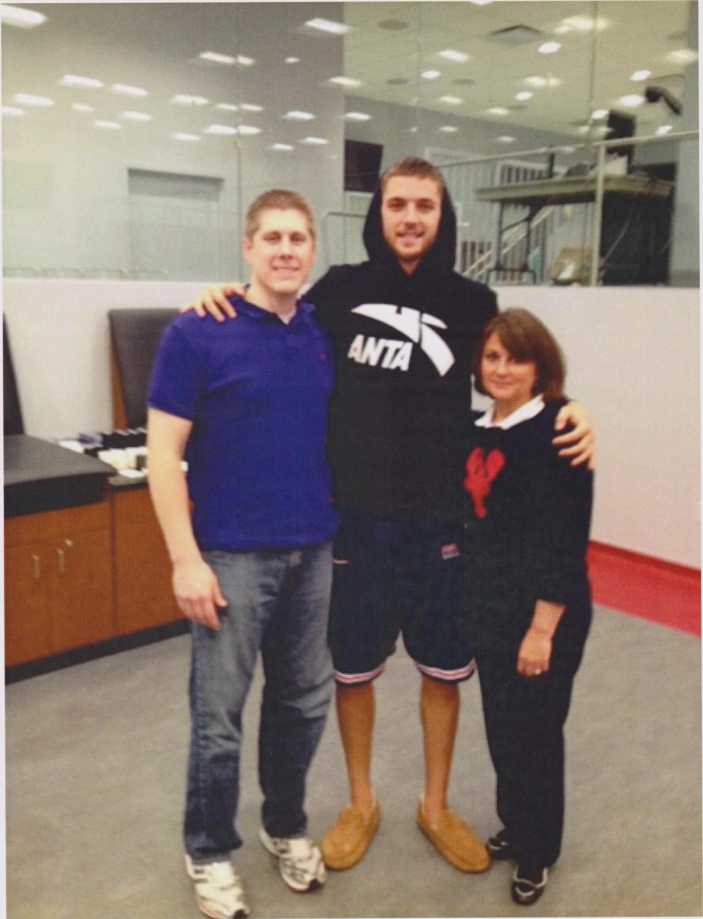 Three people are standing together in a picture.  the picture is taken in the Houston Rockets treatment room, with Chandler Parsons standing in the center of the room wearing a black hoodie, black long shorts and brown sandals.  