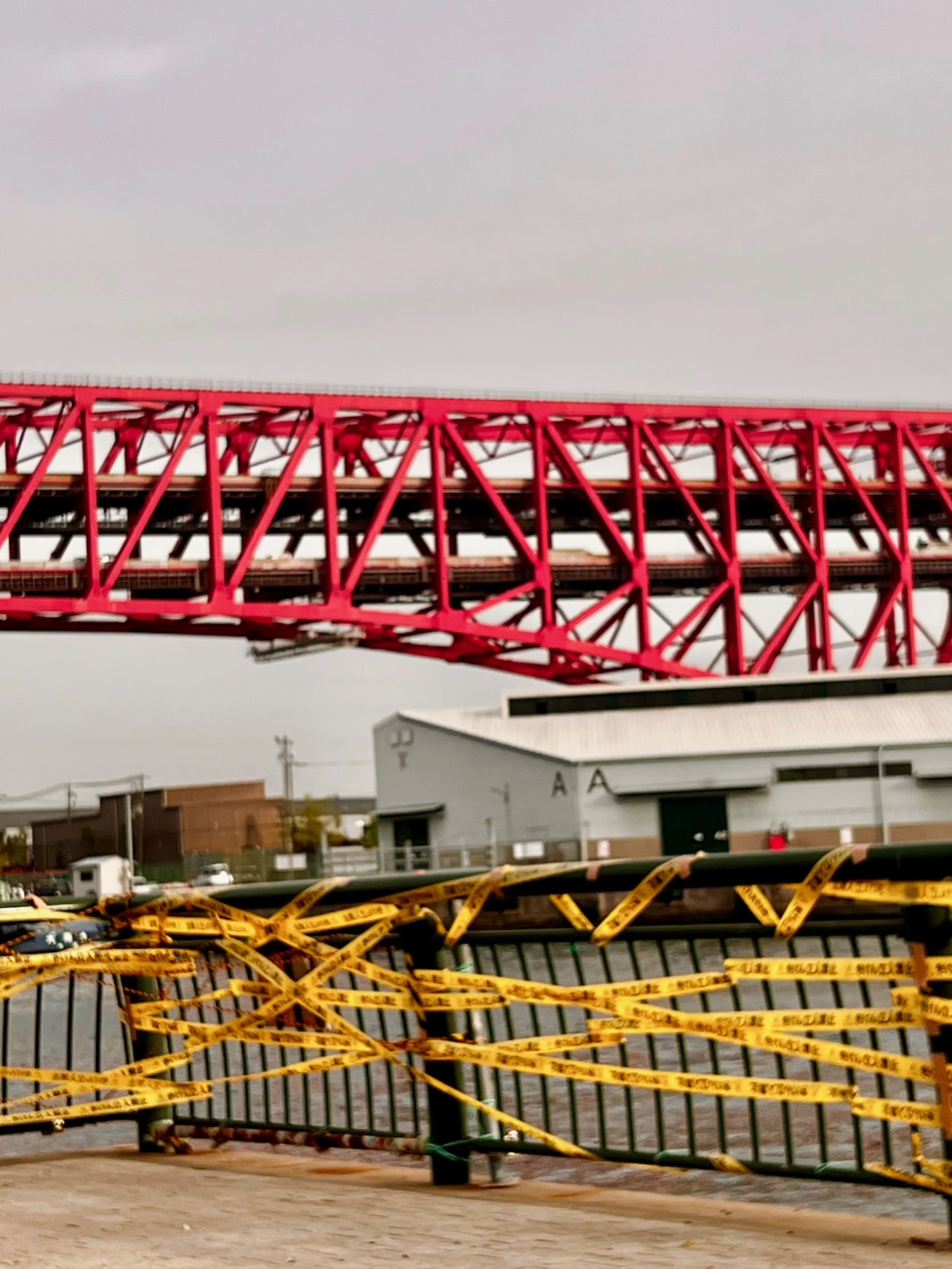 A view of a red bridge with train tracks, with yellow police tape blocking a walkway in the foreground and a gray industrial building in the background. Osaka
