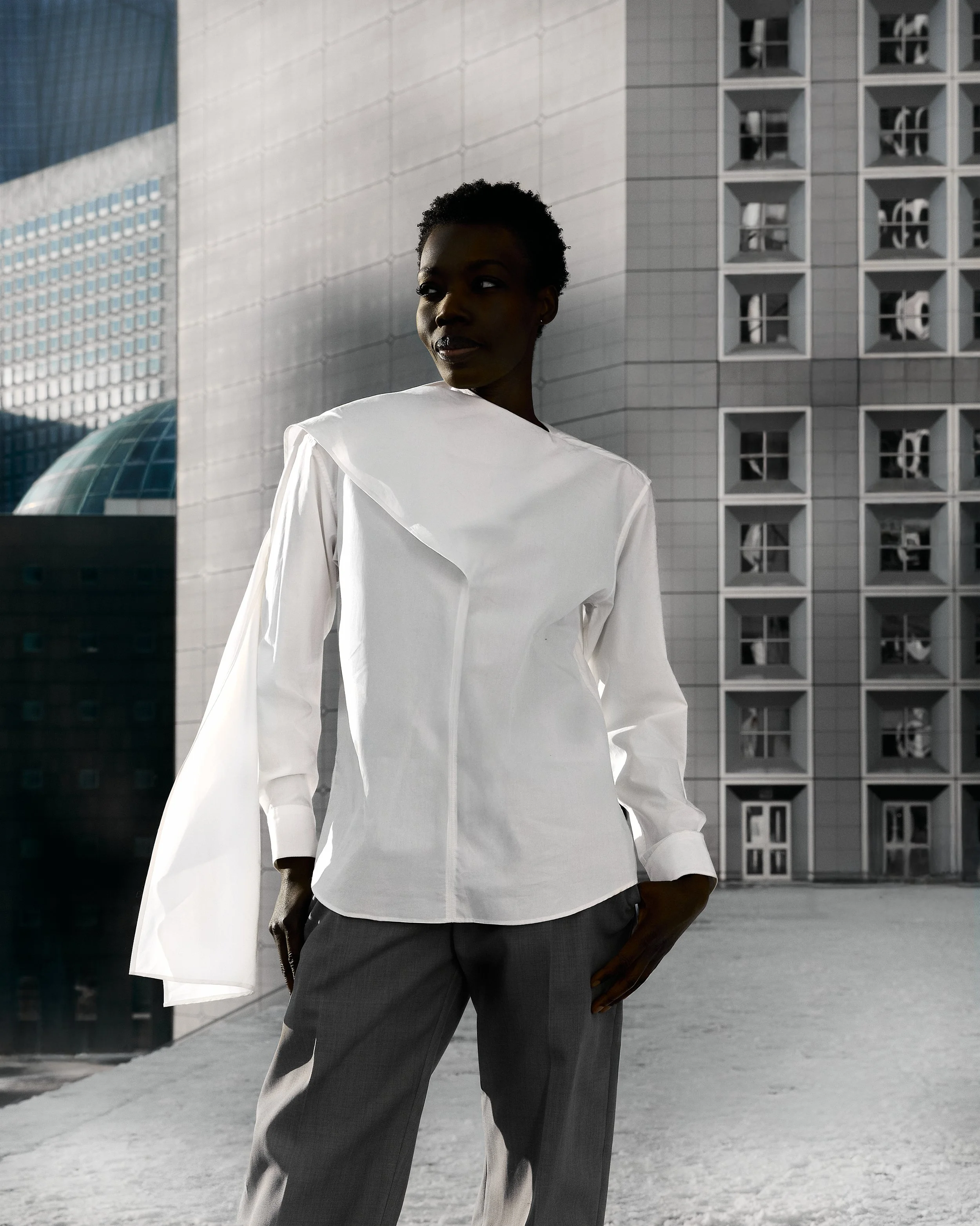 A woman with dark skin and short curly hair stands outdoors in front of modern buildings, wearing a white long-sleeve shirt and gray pants.