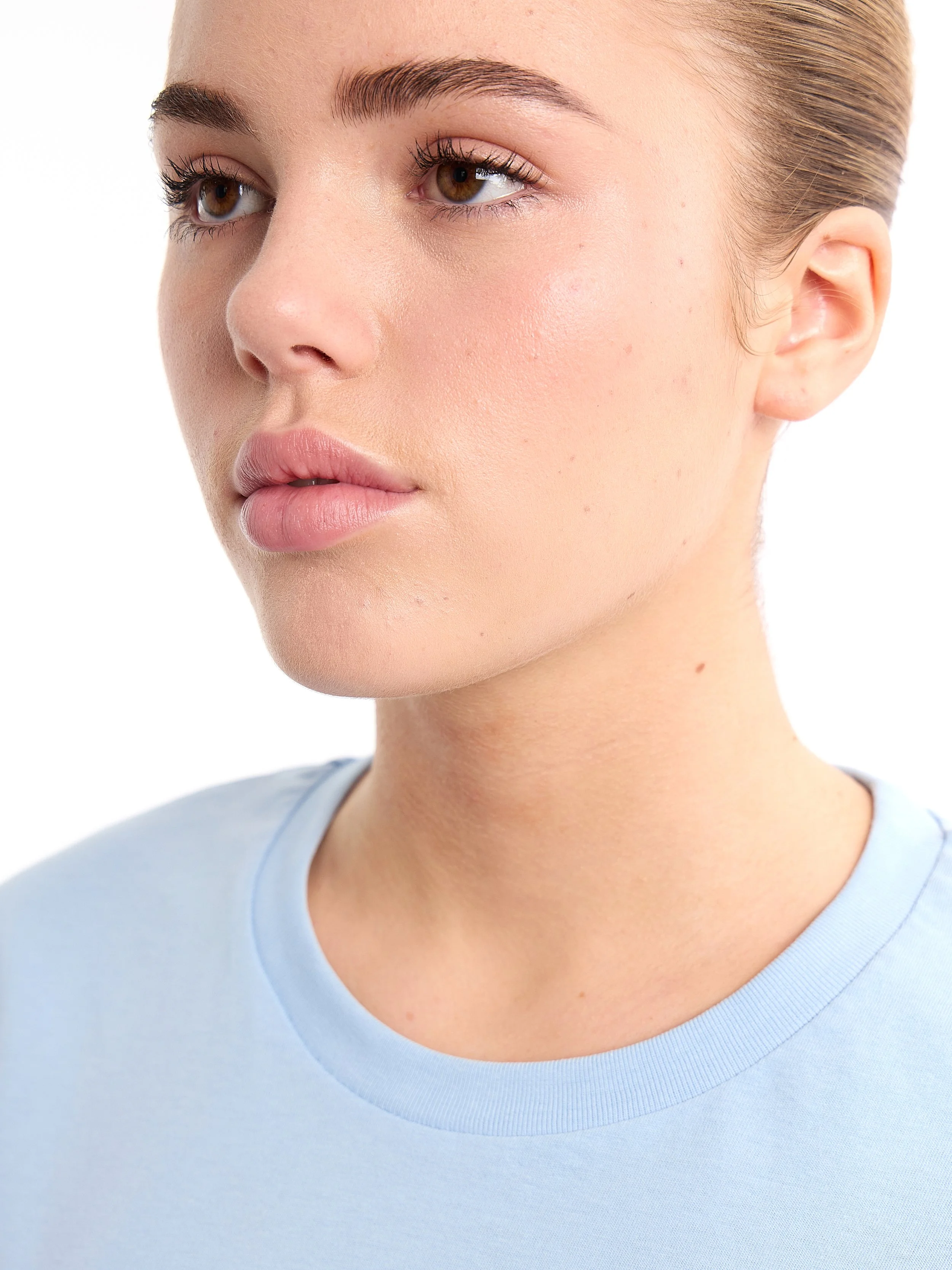 Close-up of a young woman with clear skin and light makeup, wearing a light blue shirt, against a white background. m^