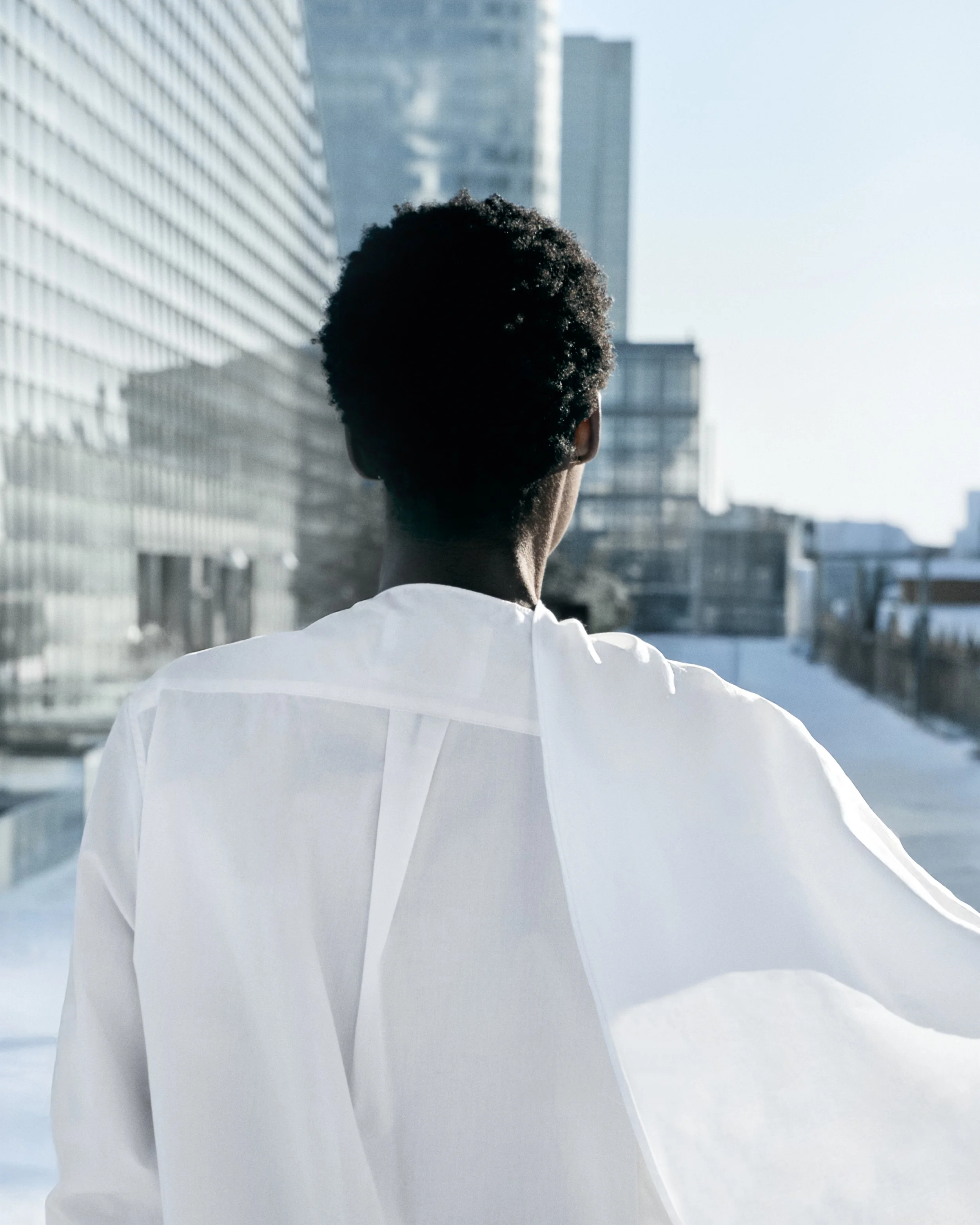 Back of a person with short, curly hair wearing a white shirt, standing outdoors in a cityscape with tall modern buildings.