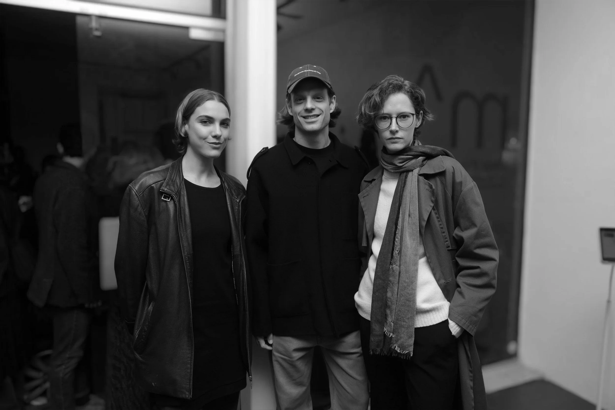 Black and white photo of three young adults standing together indoors, smiling at the camera. The person on the left is wearing a leather jacket, the middle person is wearing a cap and a dark jacket, and the person on the right has glasses, a scarf, and a jacket.