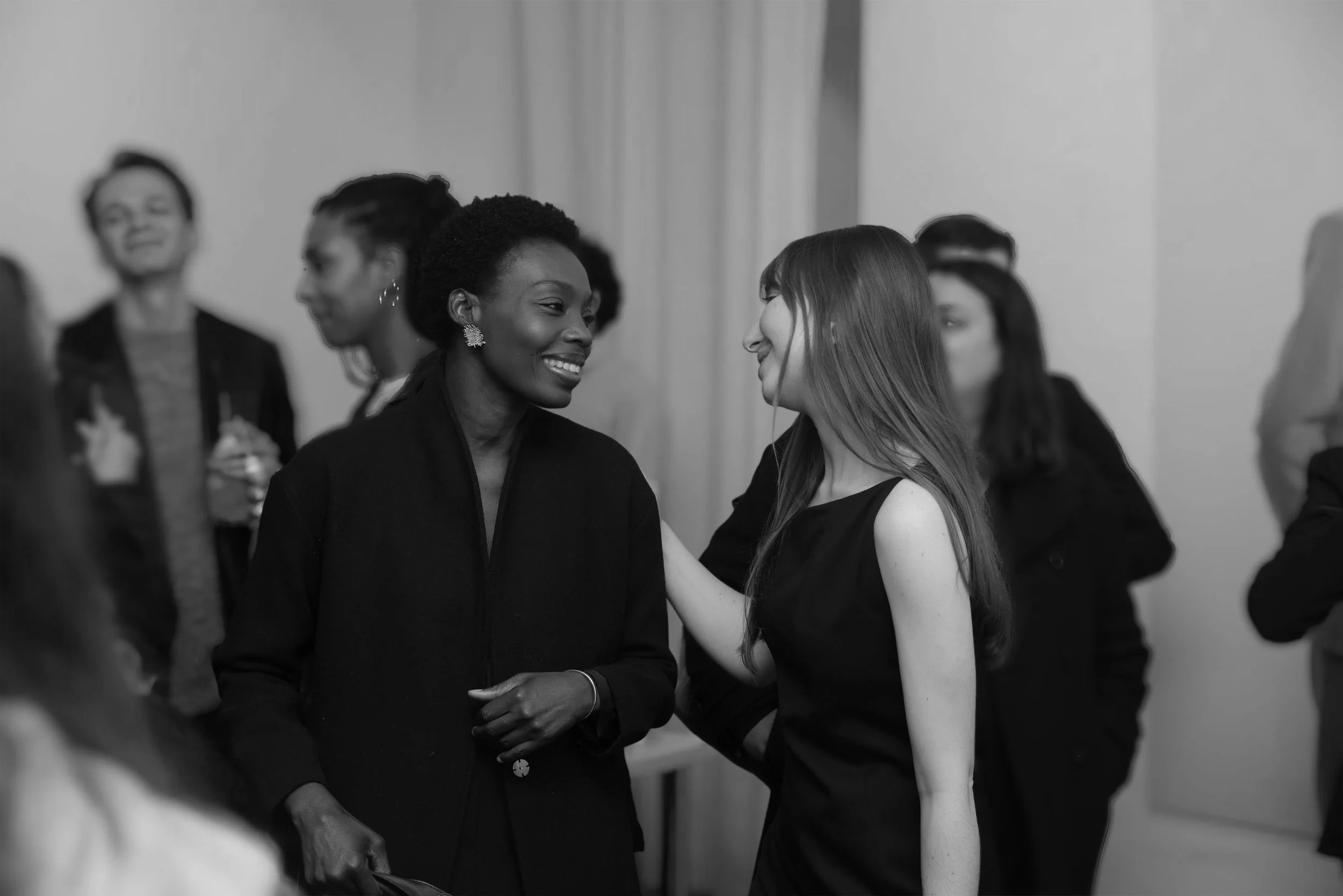 Two women smiling and talking closely at a social gathering, with other people in the background.