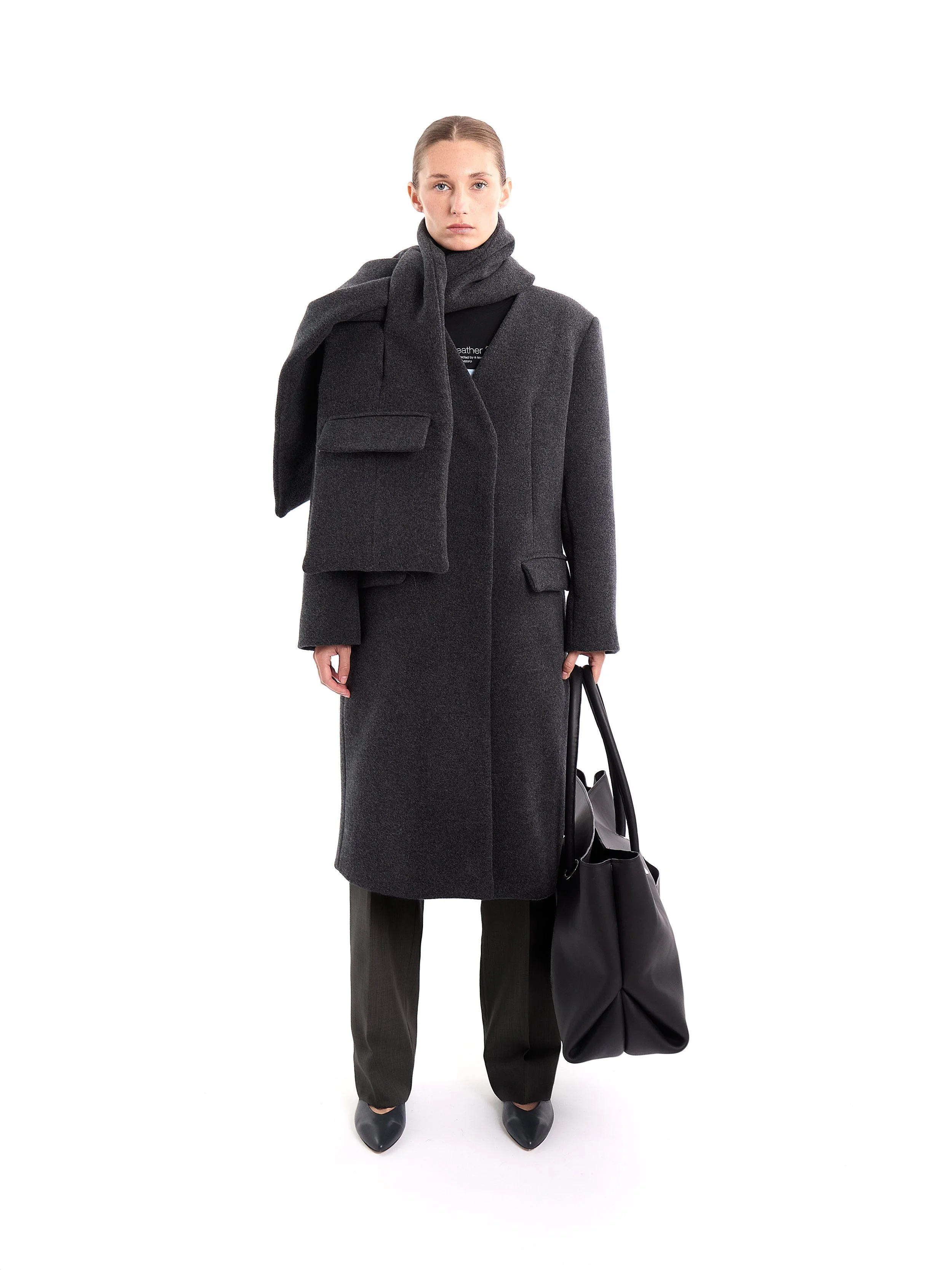 A woman in a dark gray coat and matching scarf holding a large black bag, standing against a plain white background. m^