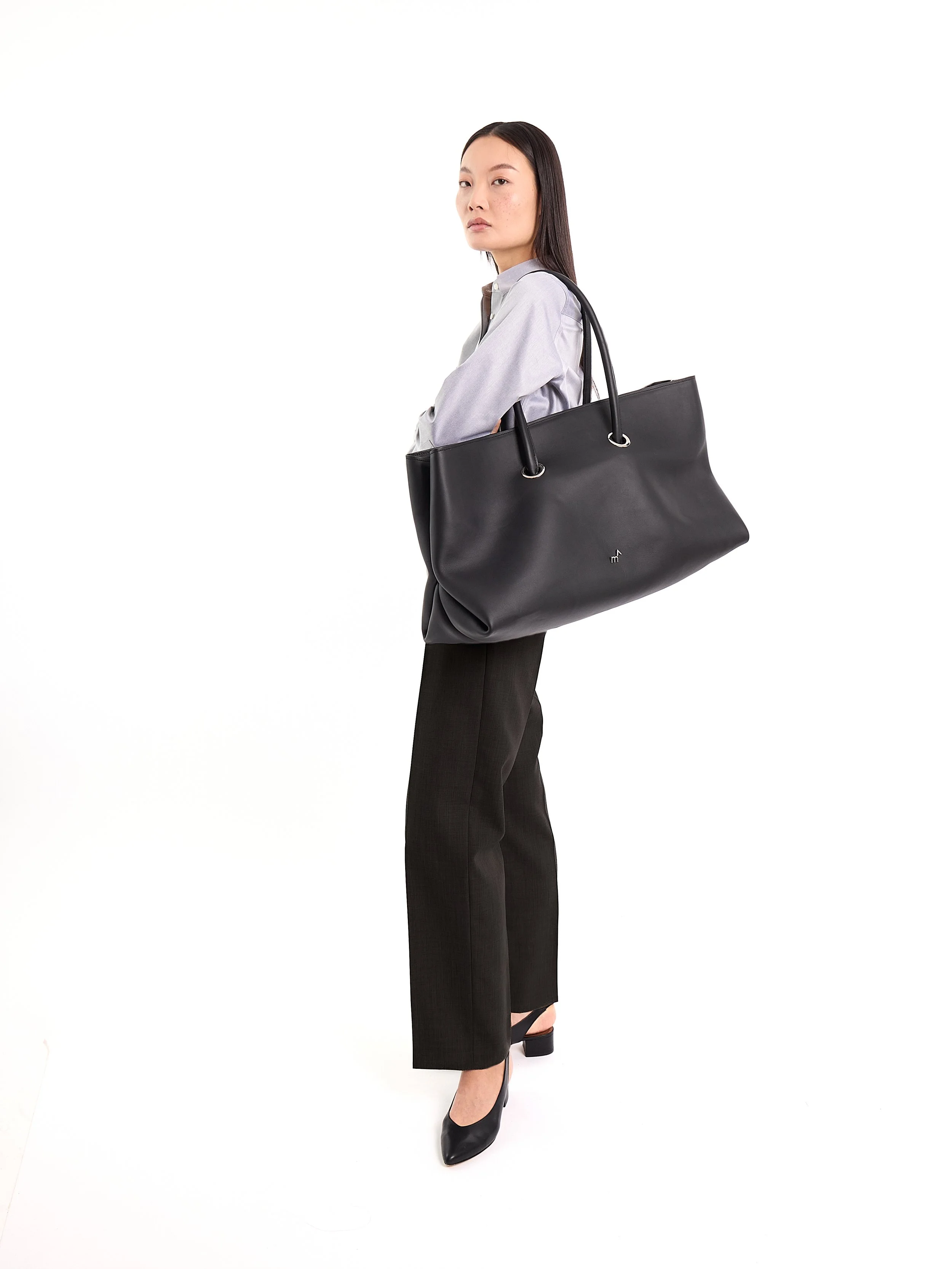 A woman stands with her arms crossed, carrying a large black tote bag over her shoulder, dressed in a light gray blouse, black wide-leg pants, and black heels, against a plain white background. m^