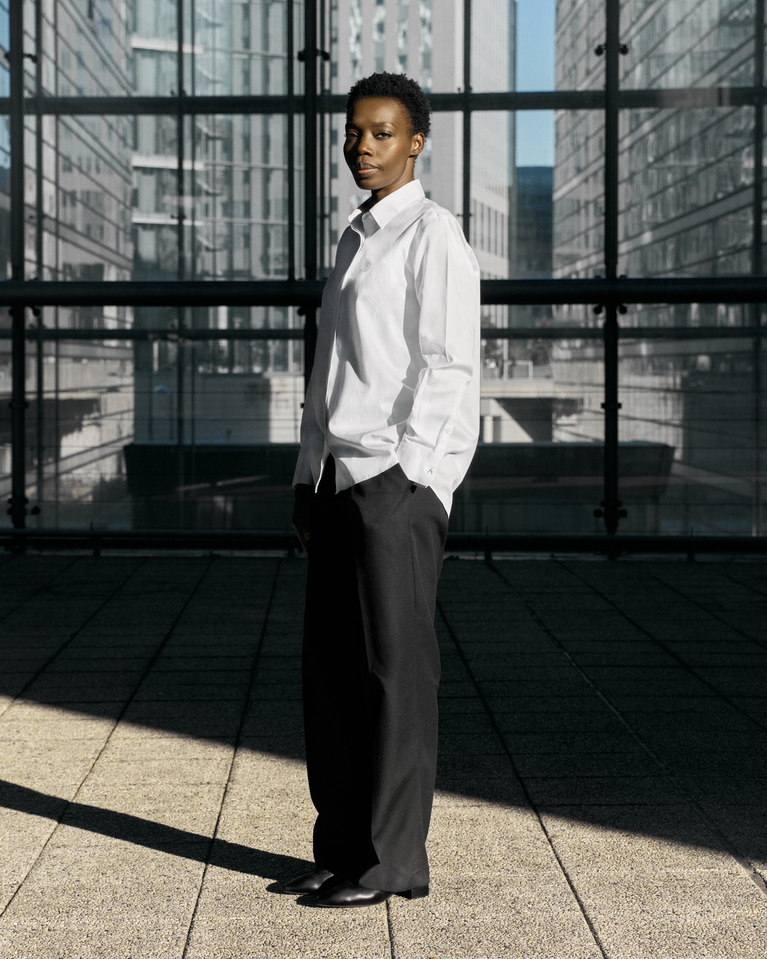 A woman stands outdoors in front of a modern glass building, wearing a white button-up shirt and black trousers, with her hands in her pockets.