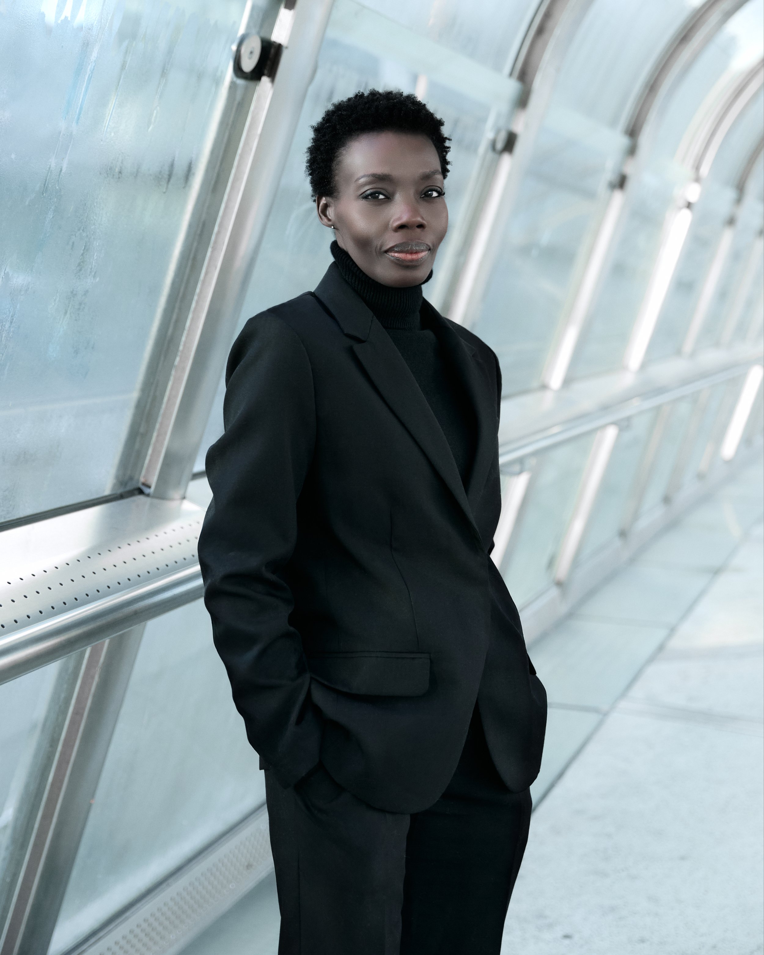 A woman wearing a black suit and turtleneck standing in a glass-covered walkway.