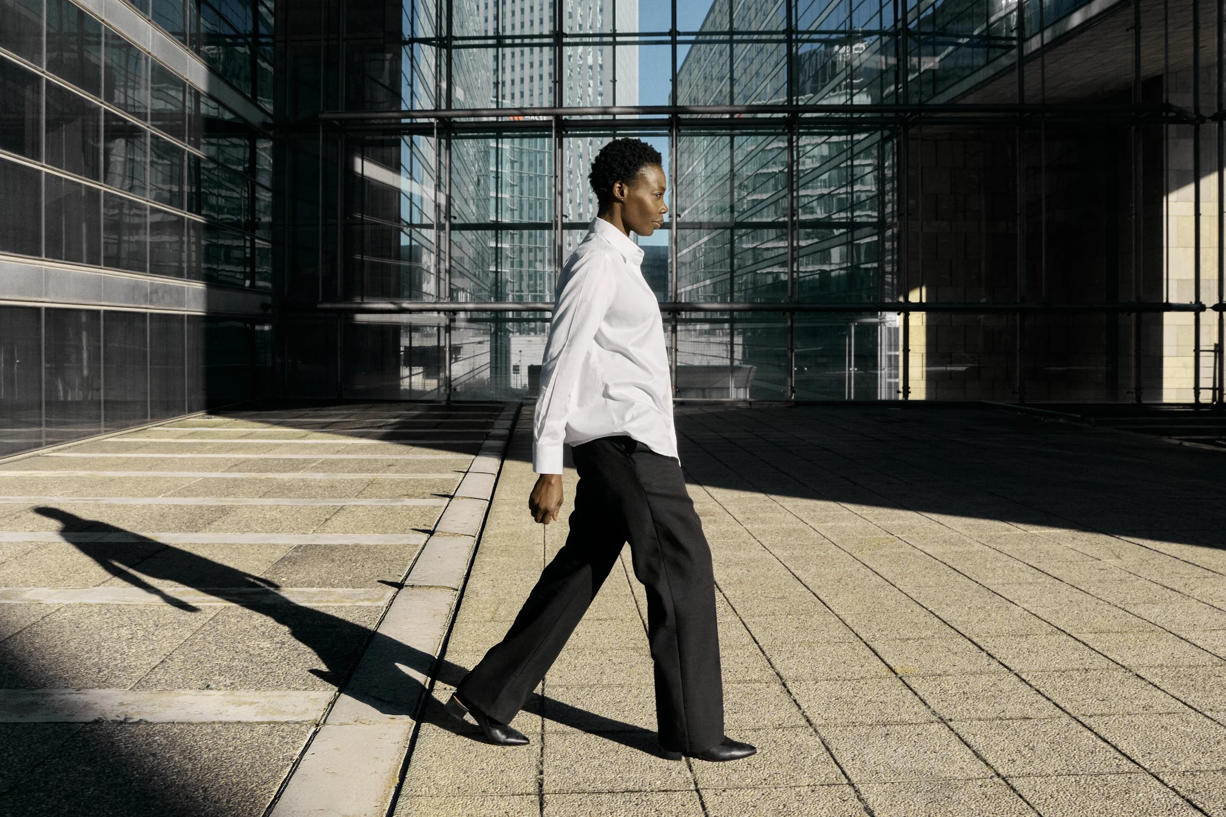 A woman walking outside a modern glass building, casting a long shadow on the pavement.
