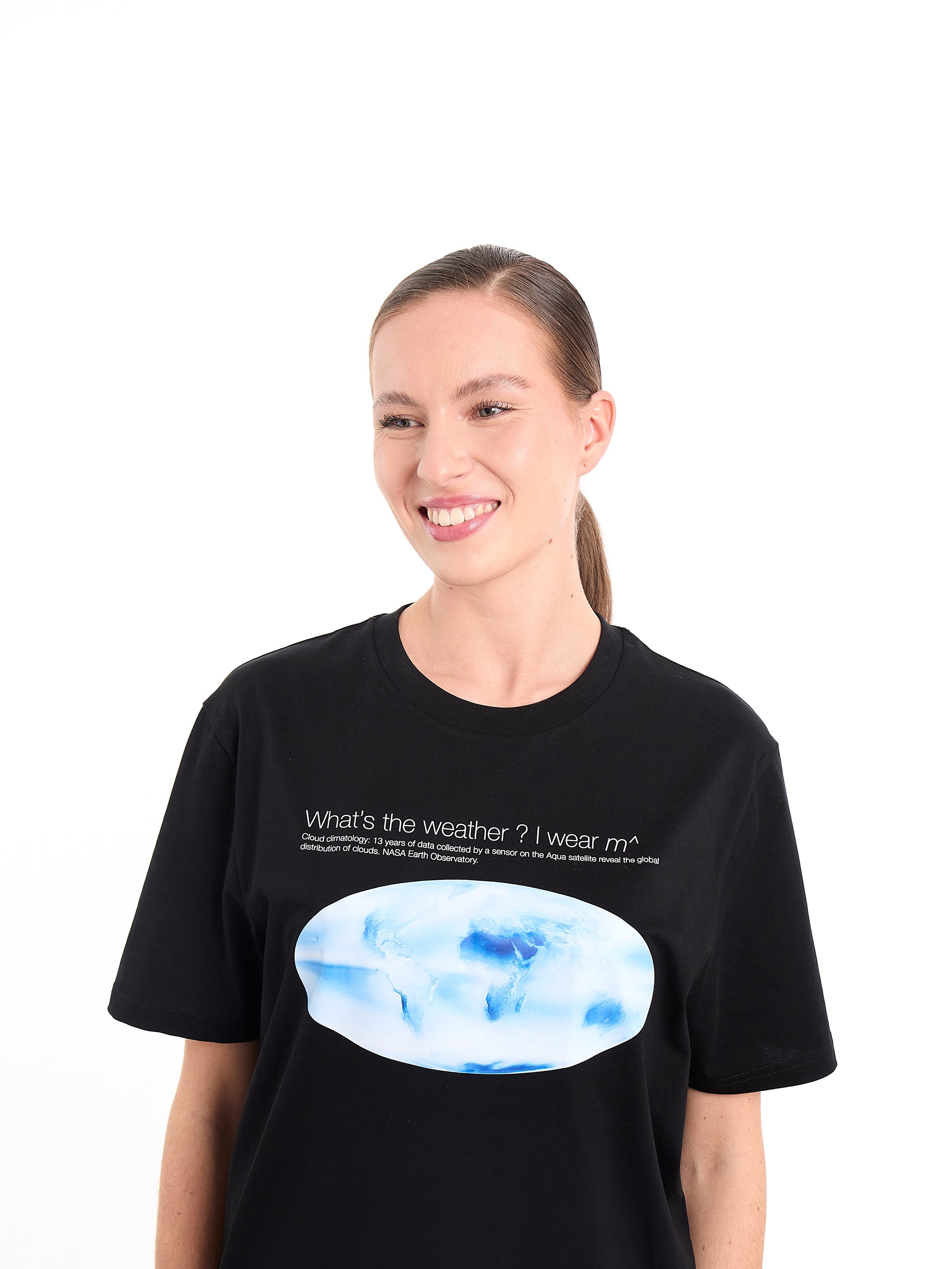 A young woman with a ponytail smiling, wearing a black T-shirt with a globe graphic and weather-related text, standing against a white background. m^