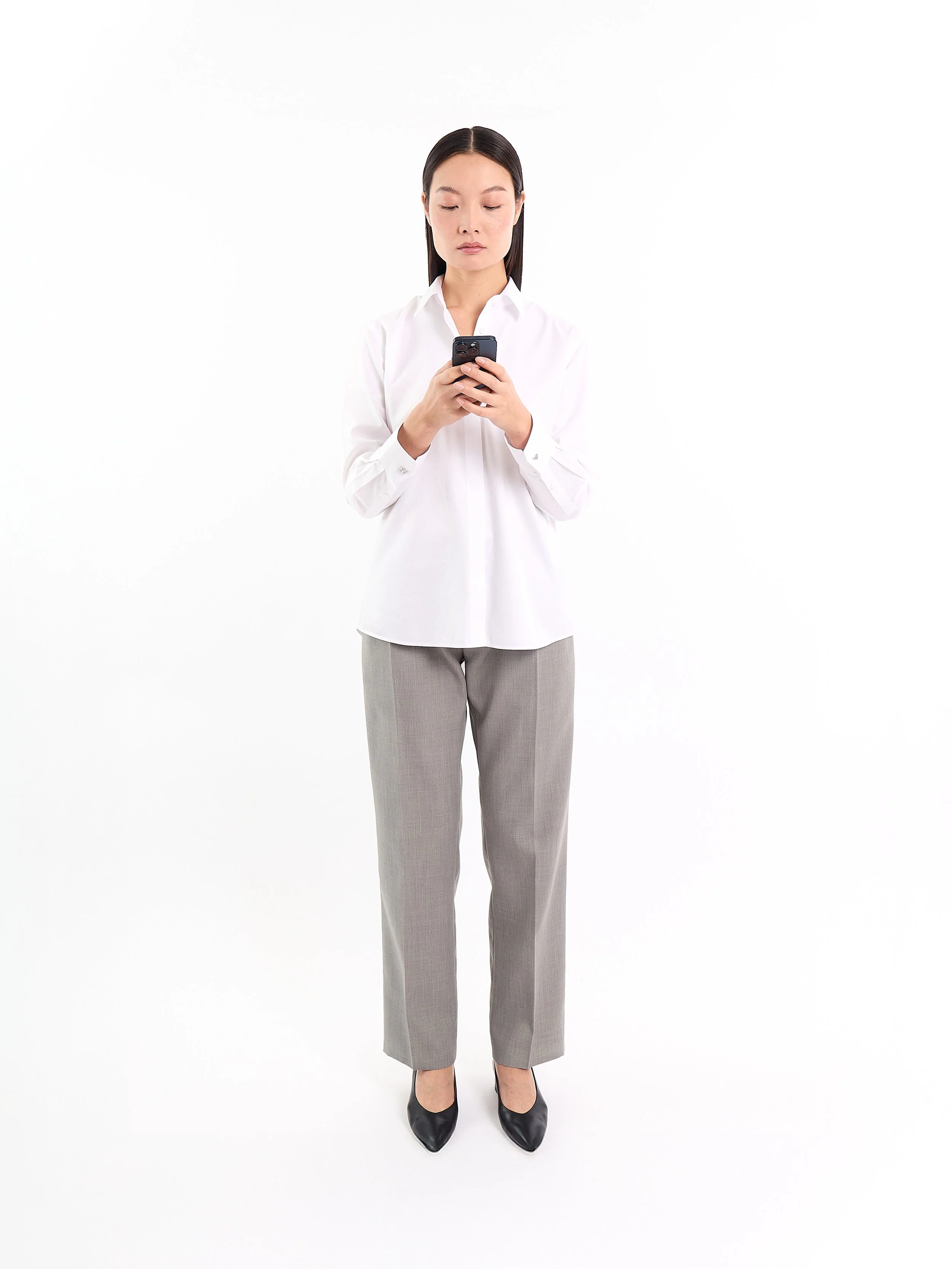 A woman standing and looking at her phone, wearing a white shirt, gray slacks, and black shoes, against a plain white background. m^