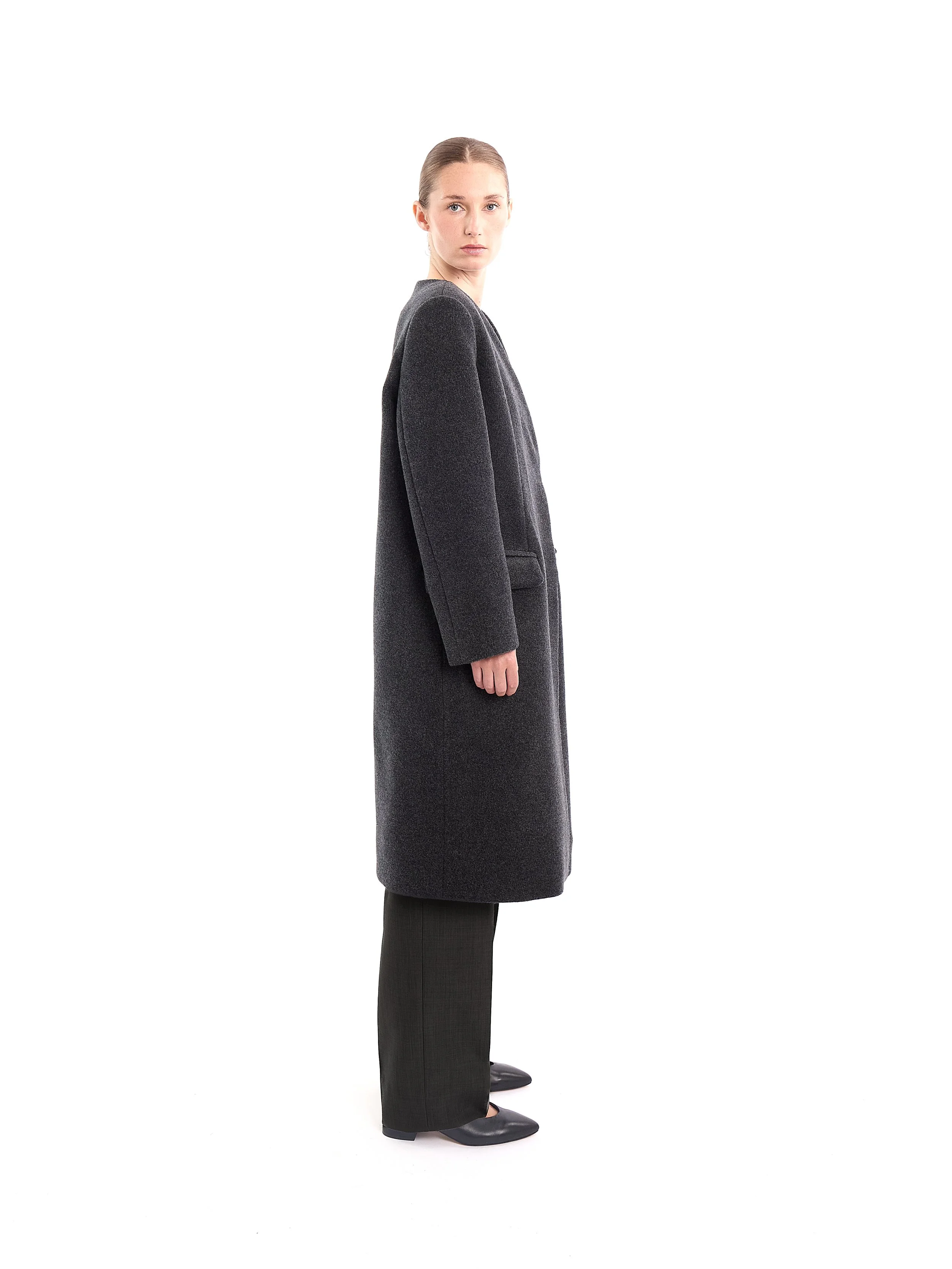 A woman wearing a long dark gray coat, black pants, and black shoes standing sideways on a white background. m^