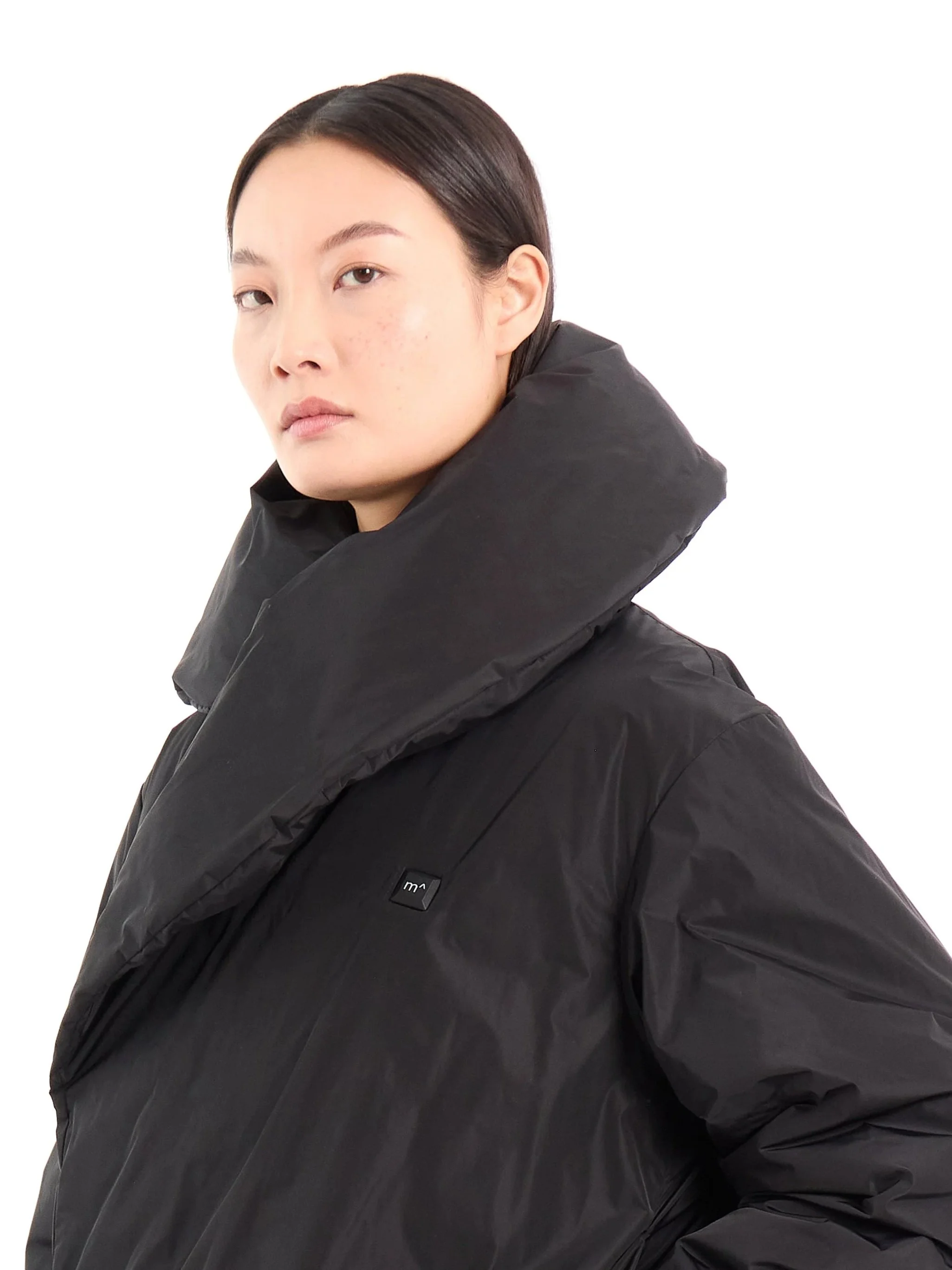 A woman with dark hair wearing a black puffer jacket with a high collar, against a plain white background. m^
