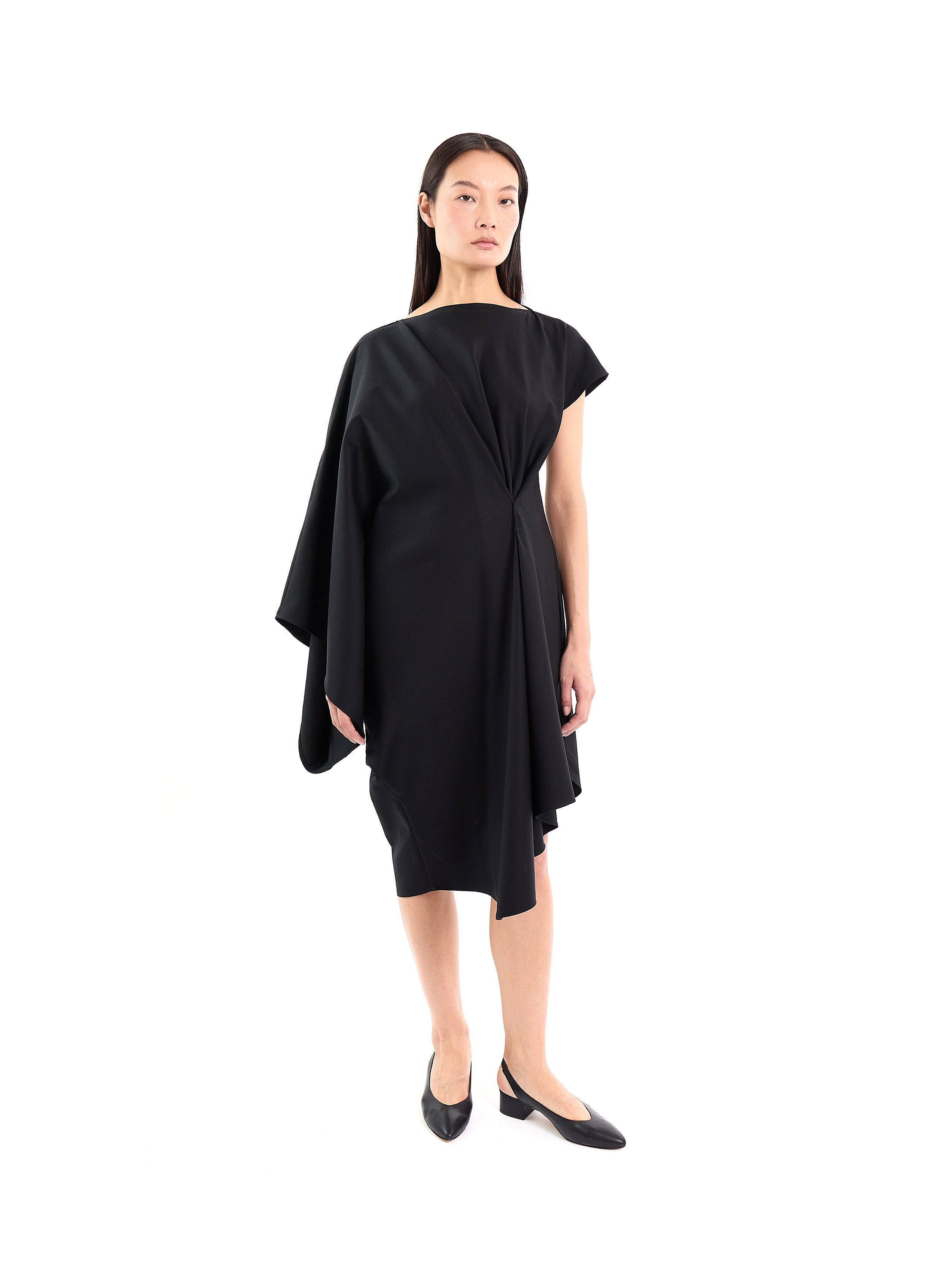 A woman with long dark hair wearing a black dress and black heels, standing against a white background. m^