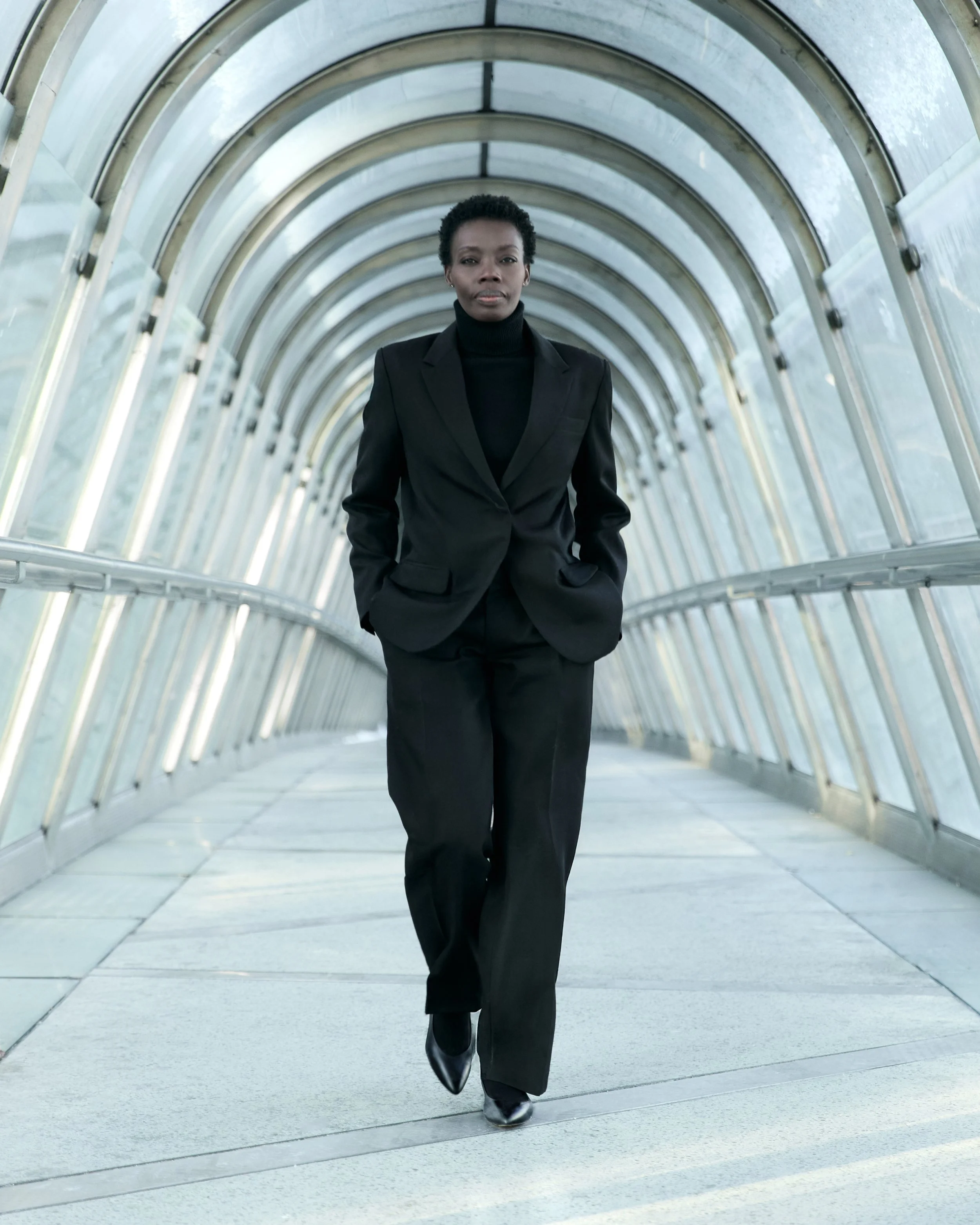 A woman in a black business suit walking through a glass-enclosed walkway with her hands in her pockets.