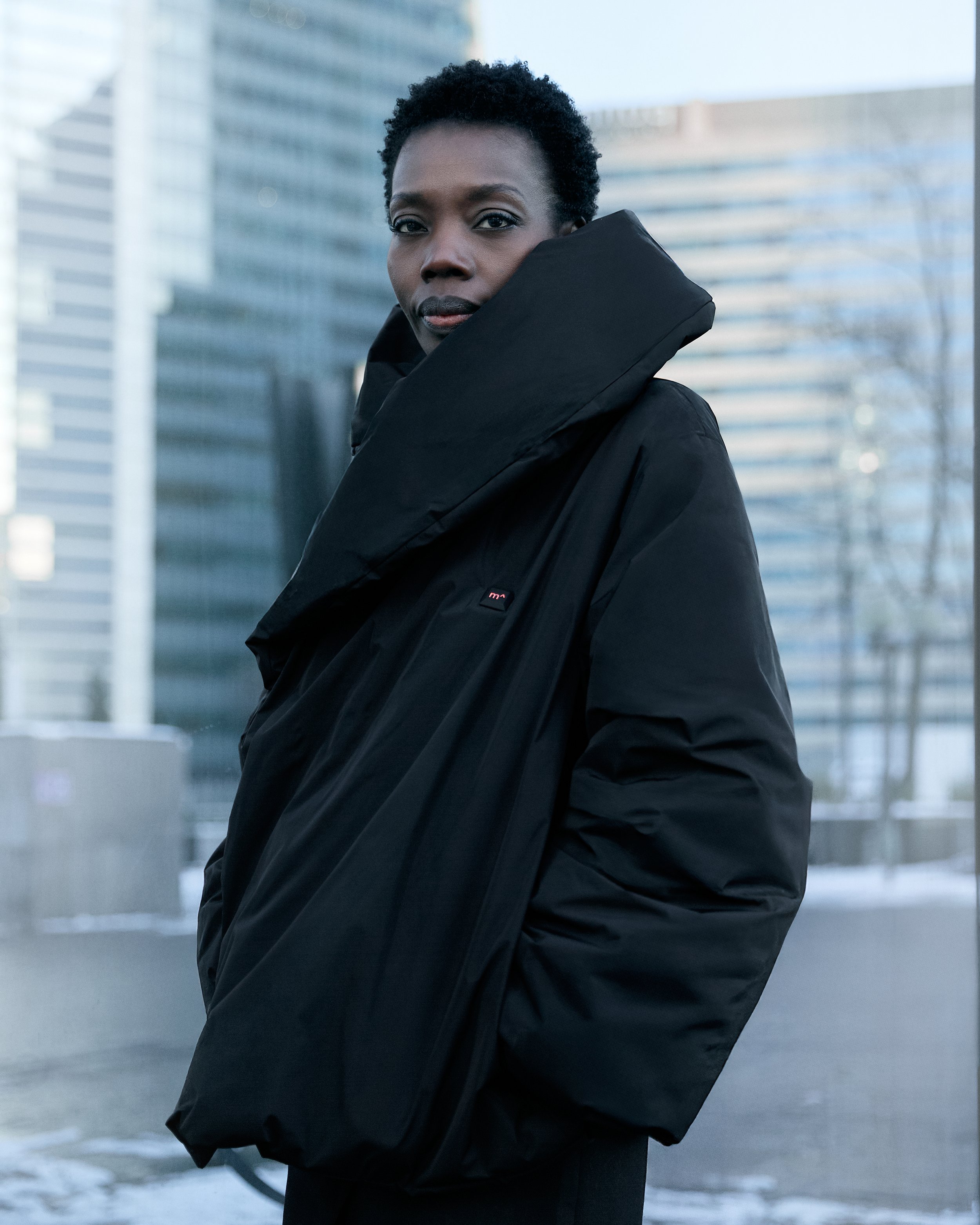 A woman with short, curly hair wearing a black jacket with a large collar, standing outdoors in front of modern office buildings. The weather appears cold.