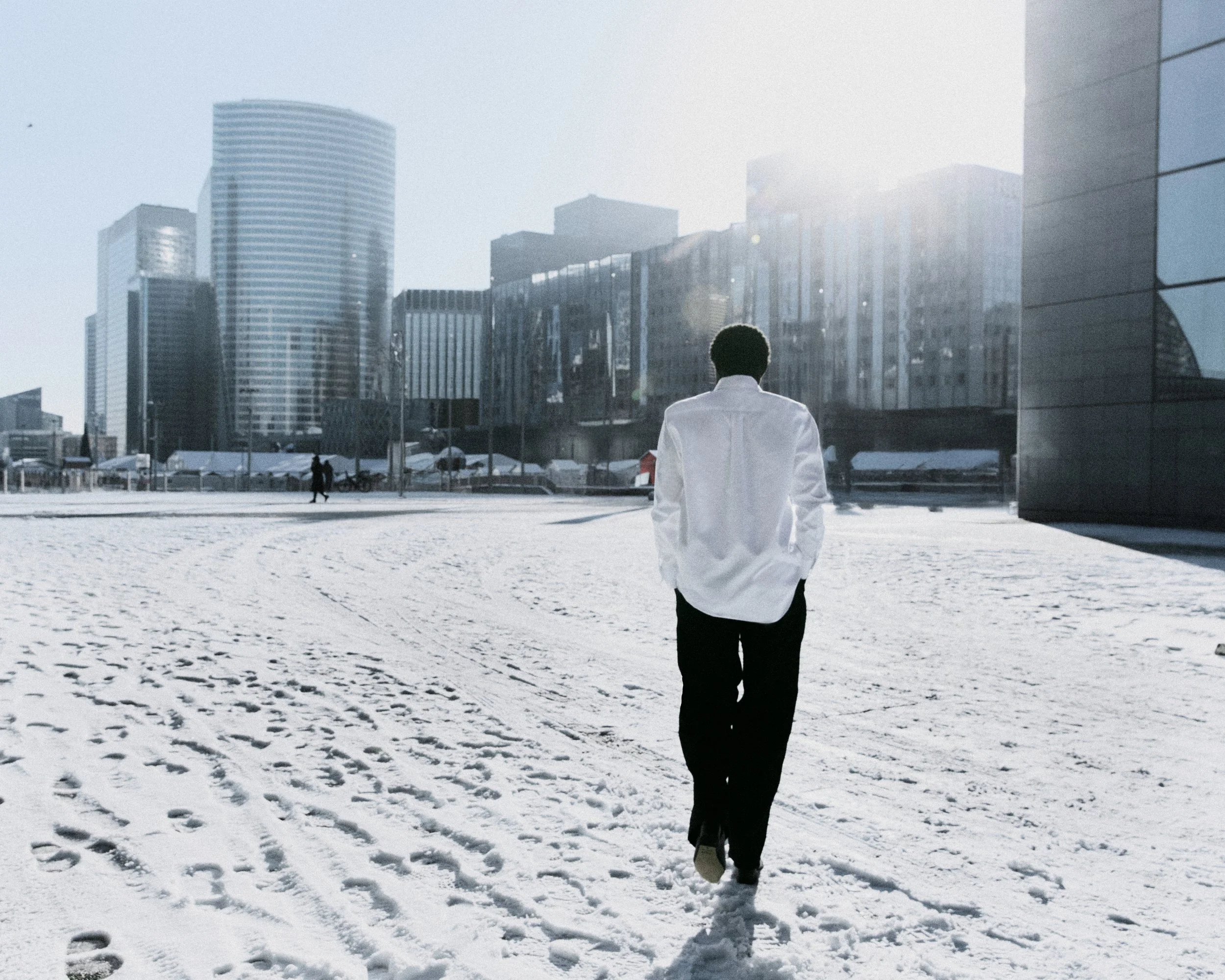 women walking on snow-covered ground in an urban cityscape with tall modern buildings, bright sunlight, and reflective windows.